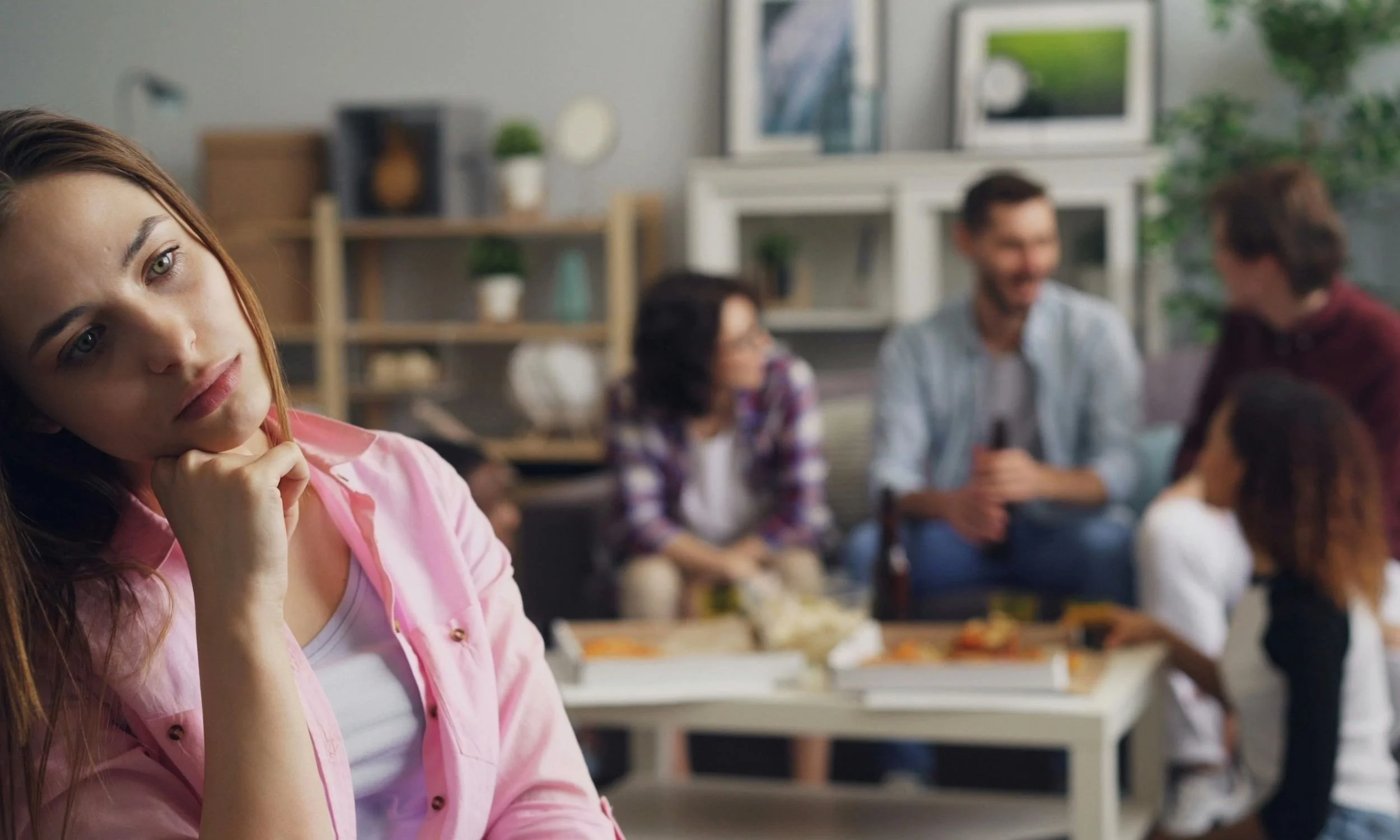 Kona situr til hliðar í félagslegum aðstæðum og virðist fjarlæg á meðan aðrir spjalla í bakgrunni.