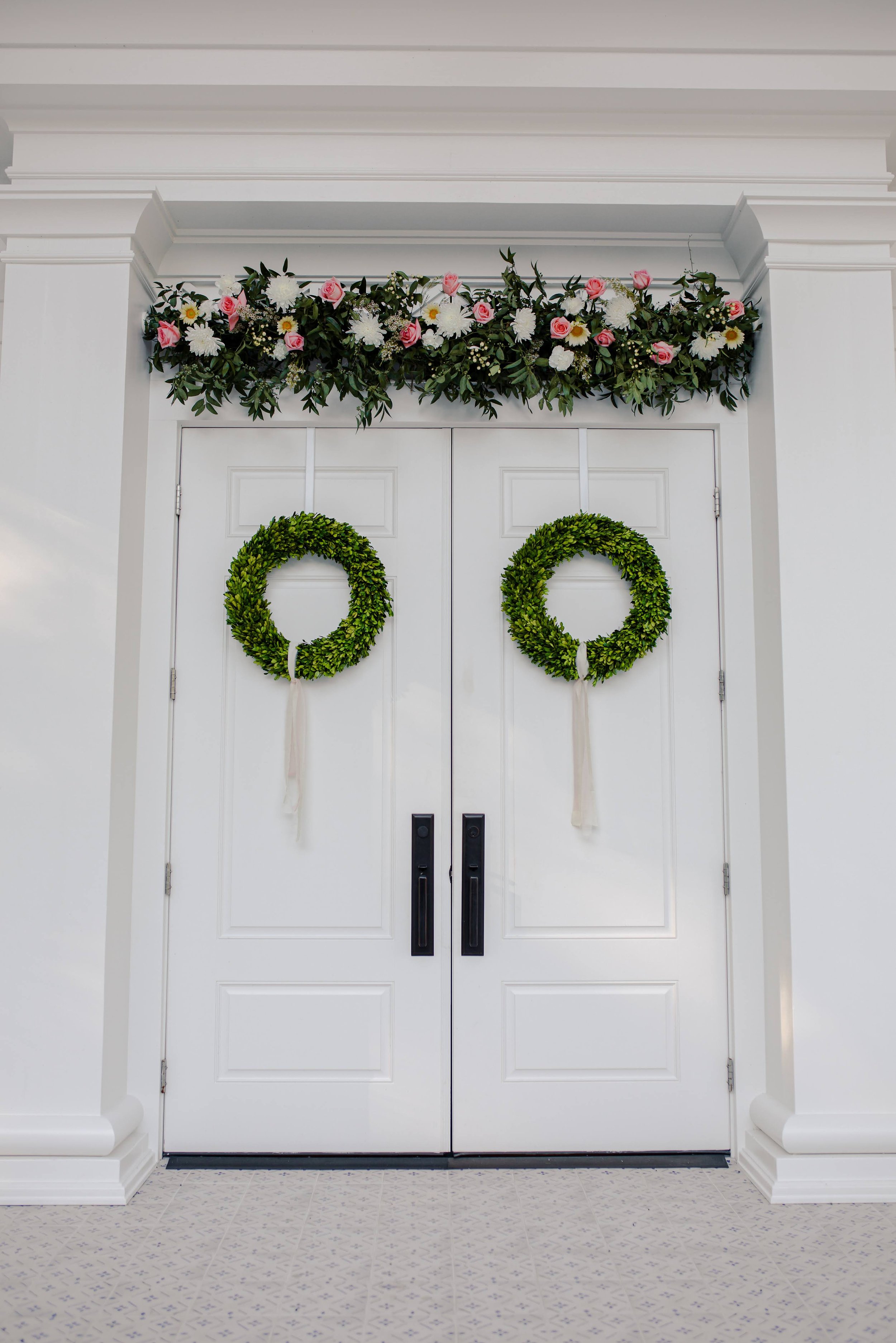 Double white doors decorated with two green wreaths hanging side by side, topped by a floral arrangement featuring pink roses, white daisies, and green foliage.