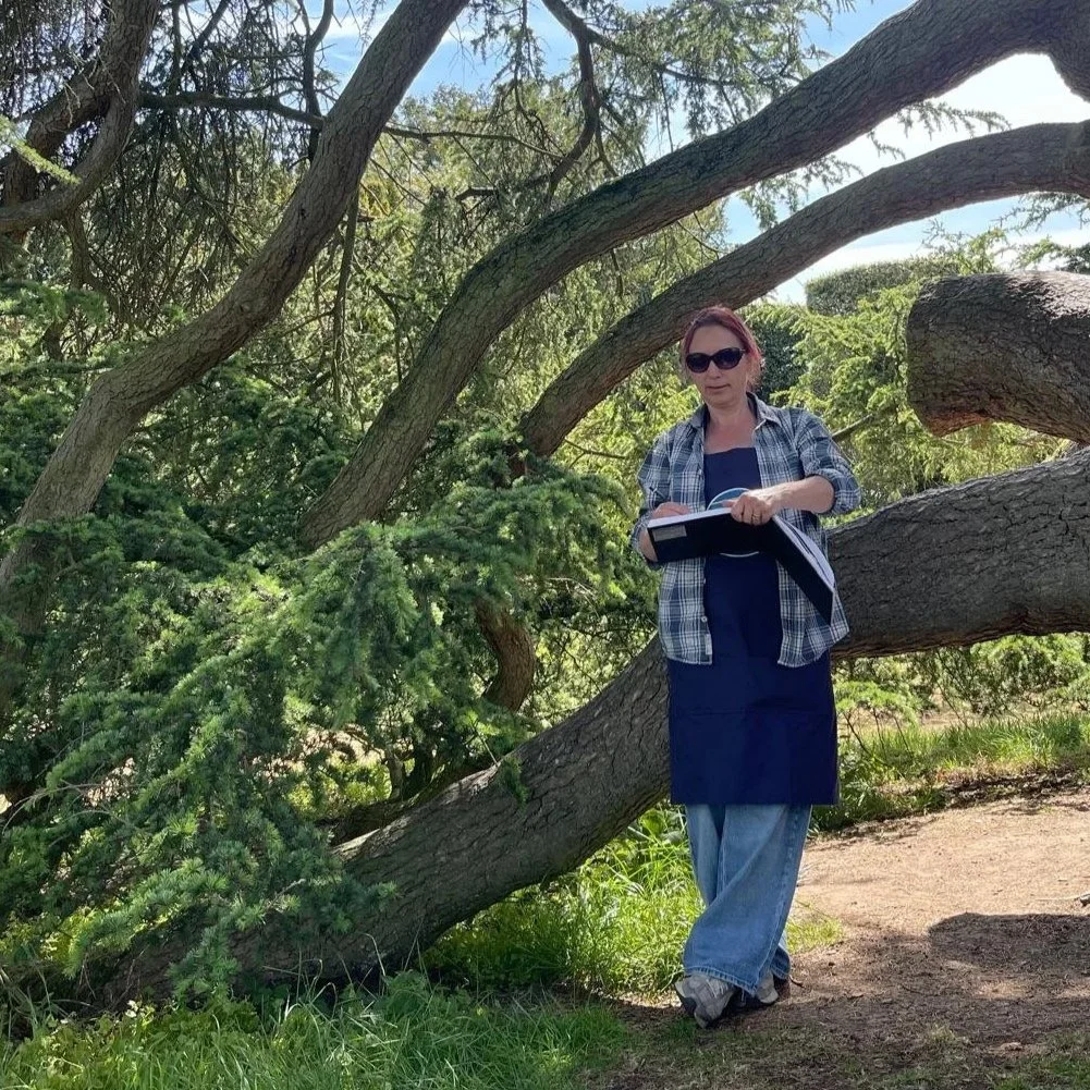 Alison Stafford Abstract Artist standing outdoors leaning on a tree, sketching, wearing sunglasses, a plaid shirt, and jeans.