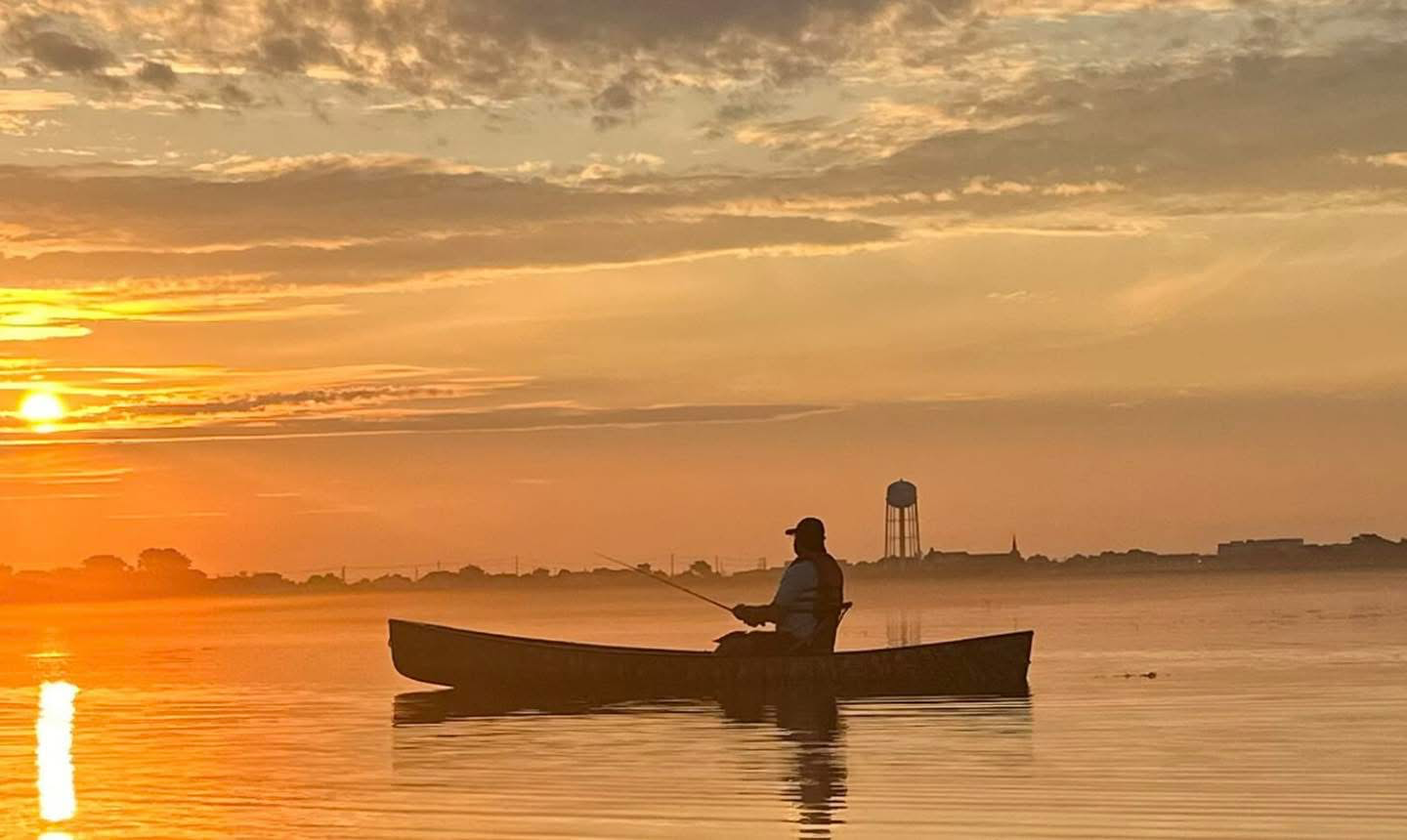 A person in a boat fishing on a calm body of water during sunrise or sunset, with a water tower and distant buildings in the background, and partly cloudy sky.