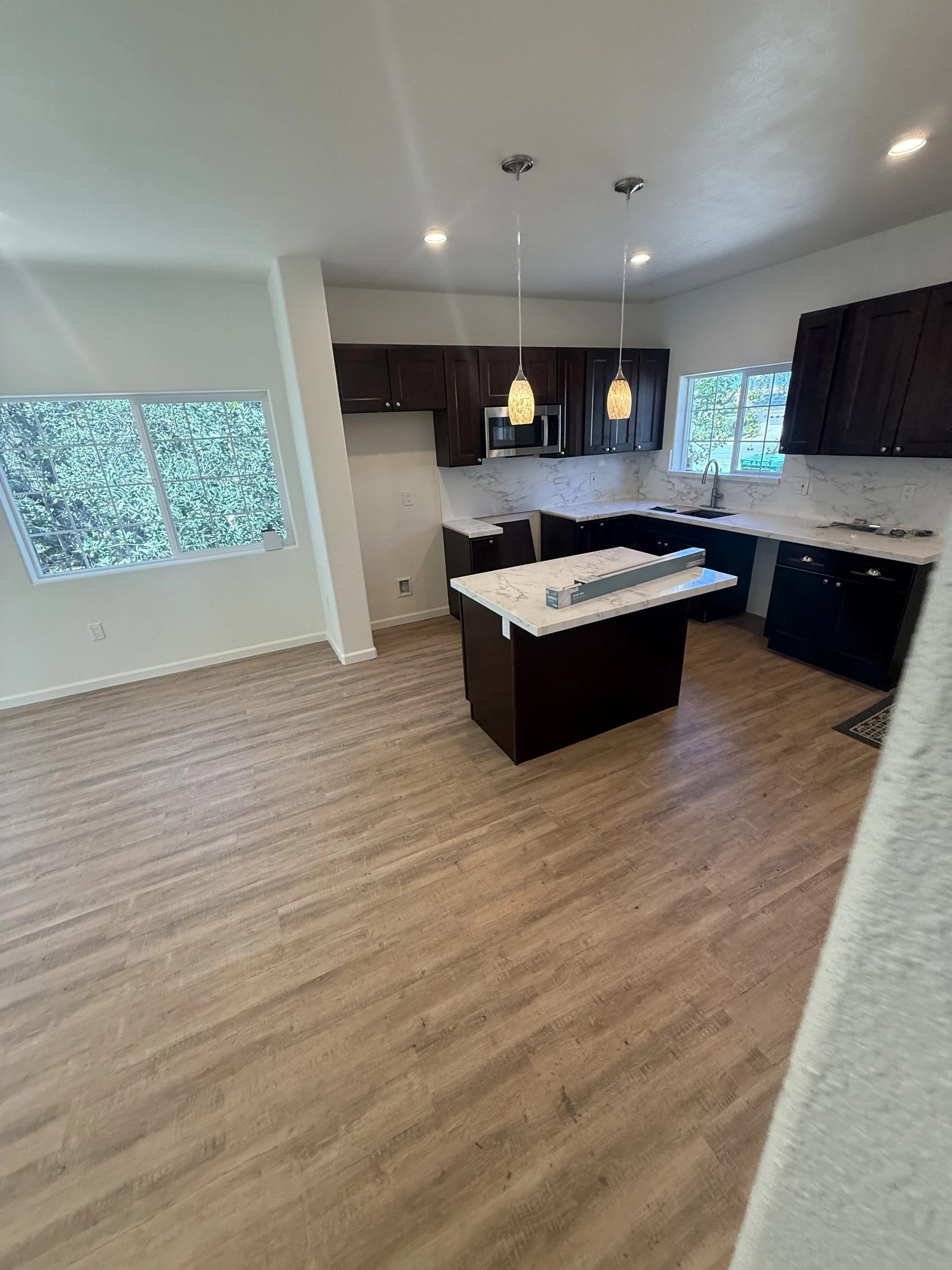 Modern kitchen with dark wooden cabinets, white marble countertops, and a small island. Large windows let in natural light, and two pendant lights hang from the ceiling.