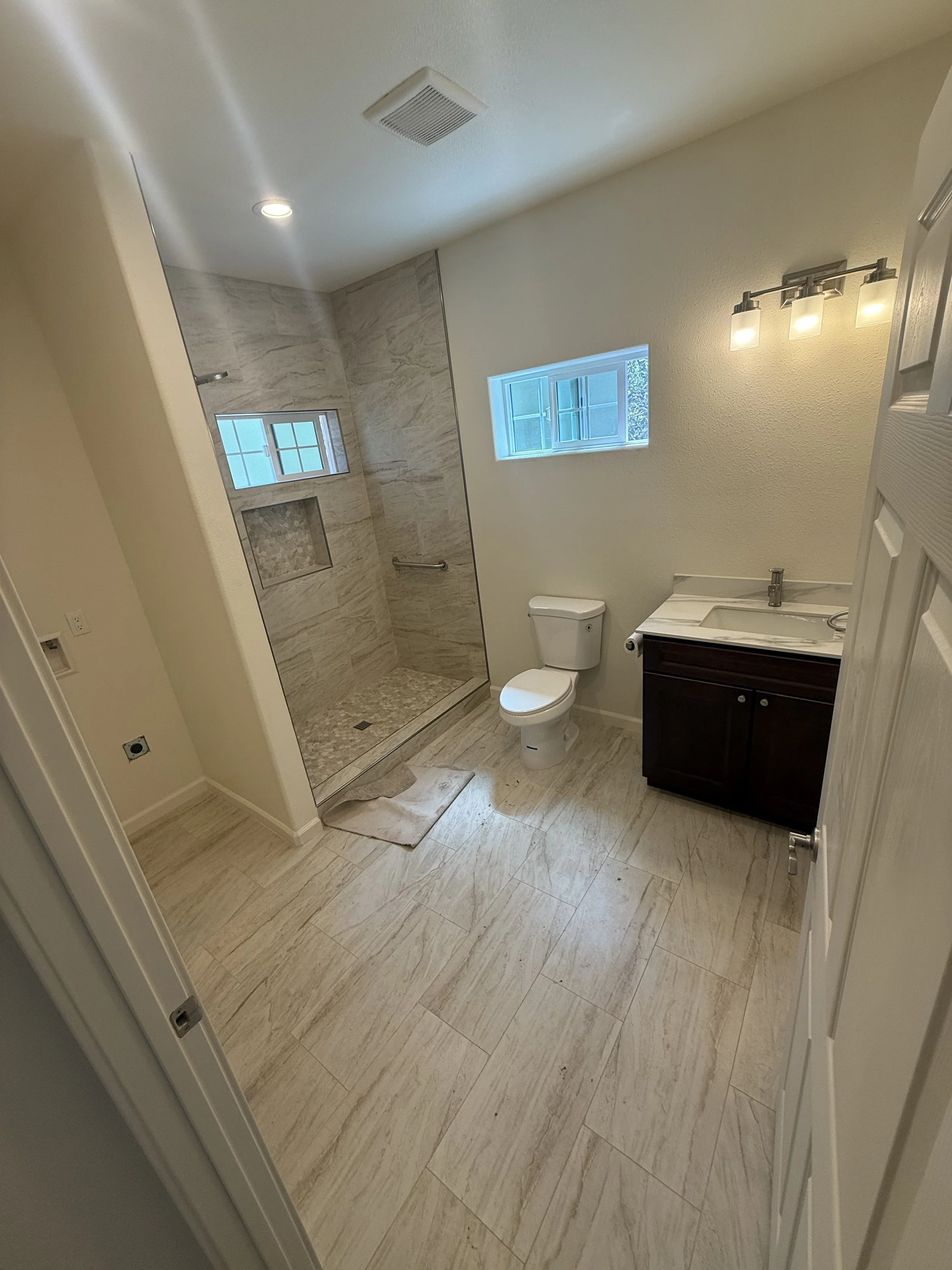 Bathroom with beige tiled floor and shower with marble walls, a small window, a toilet, and a vanity with a sink and dark cabinetry.