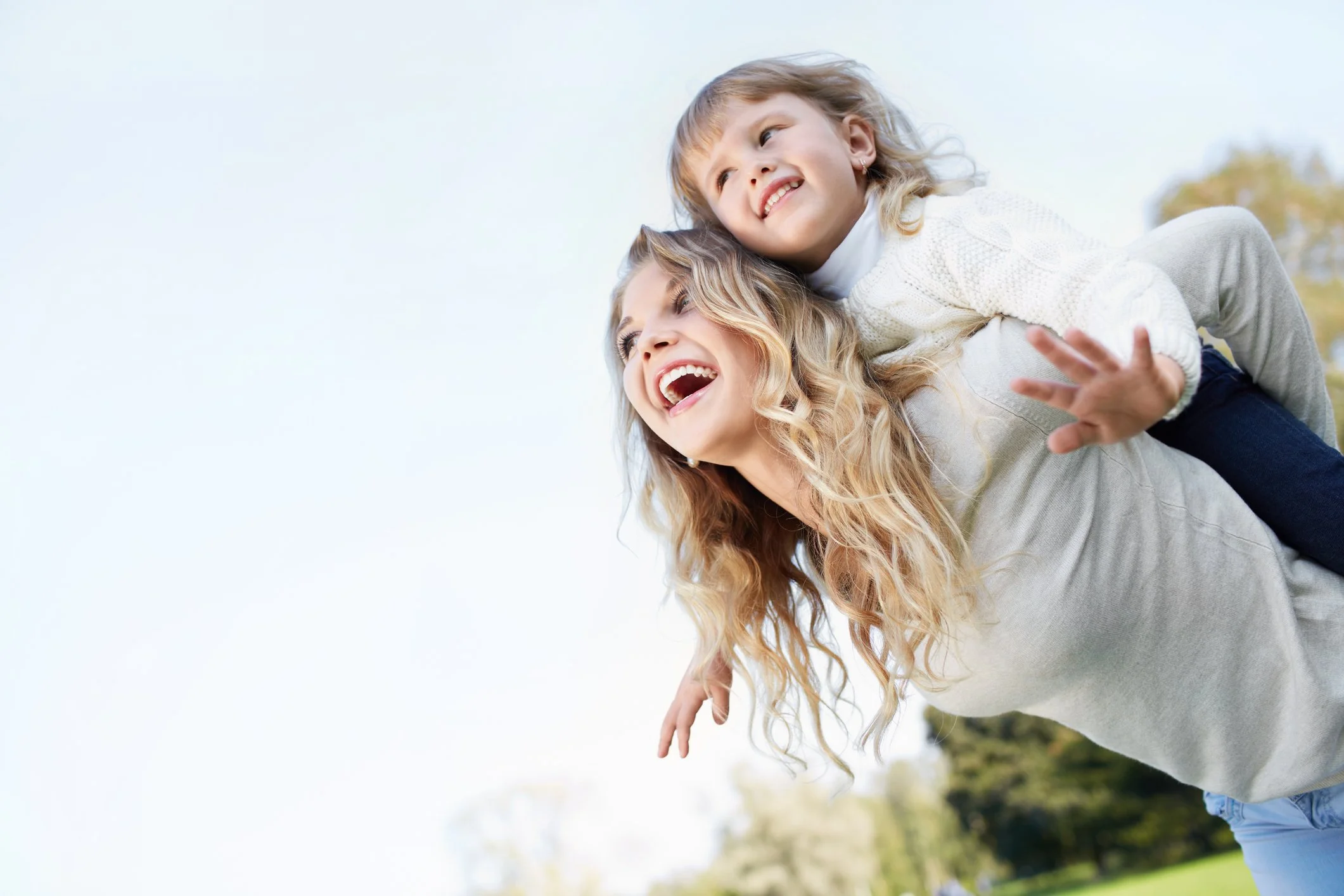 A woman with long blonde hair giving a piggyback ride to a young girl with curly hair, both smiling and enjoying outdoor activities in a park.