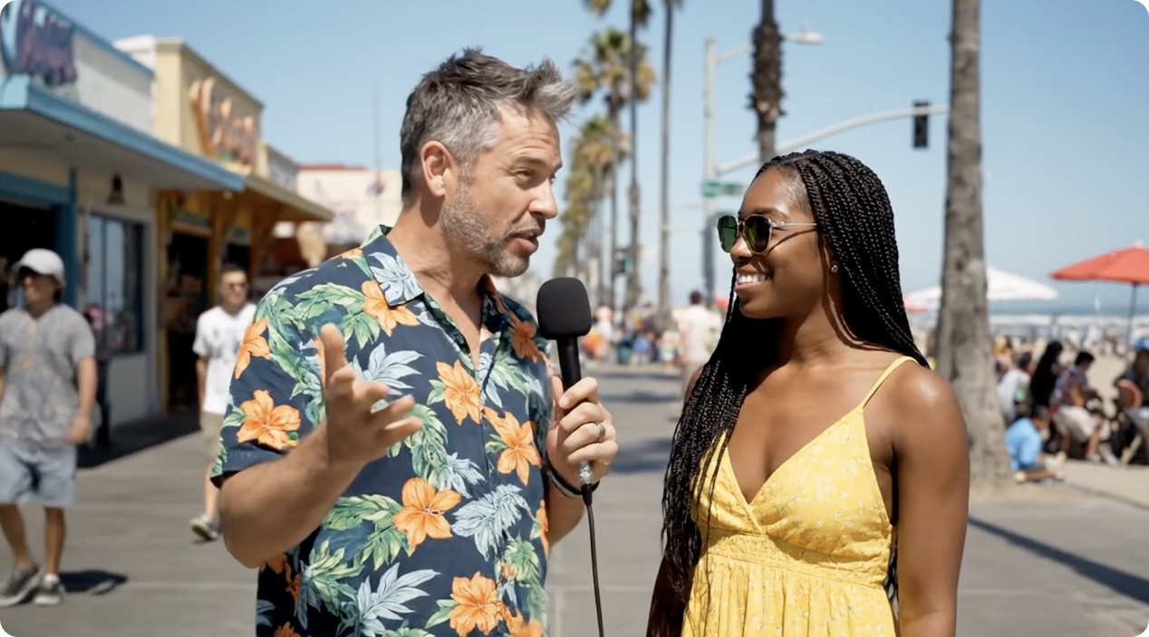 A man with gray hair and beard, wearing a floral shirt, is holding a microphone and interviewing a smiling woman with long braided hair, sunglasses, and a yellow summer dress on a sunny beachside boardwalk with palm trees, shops, and people in the background.