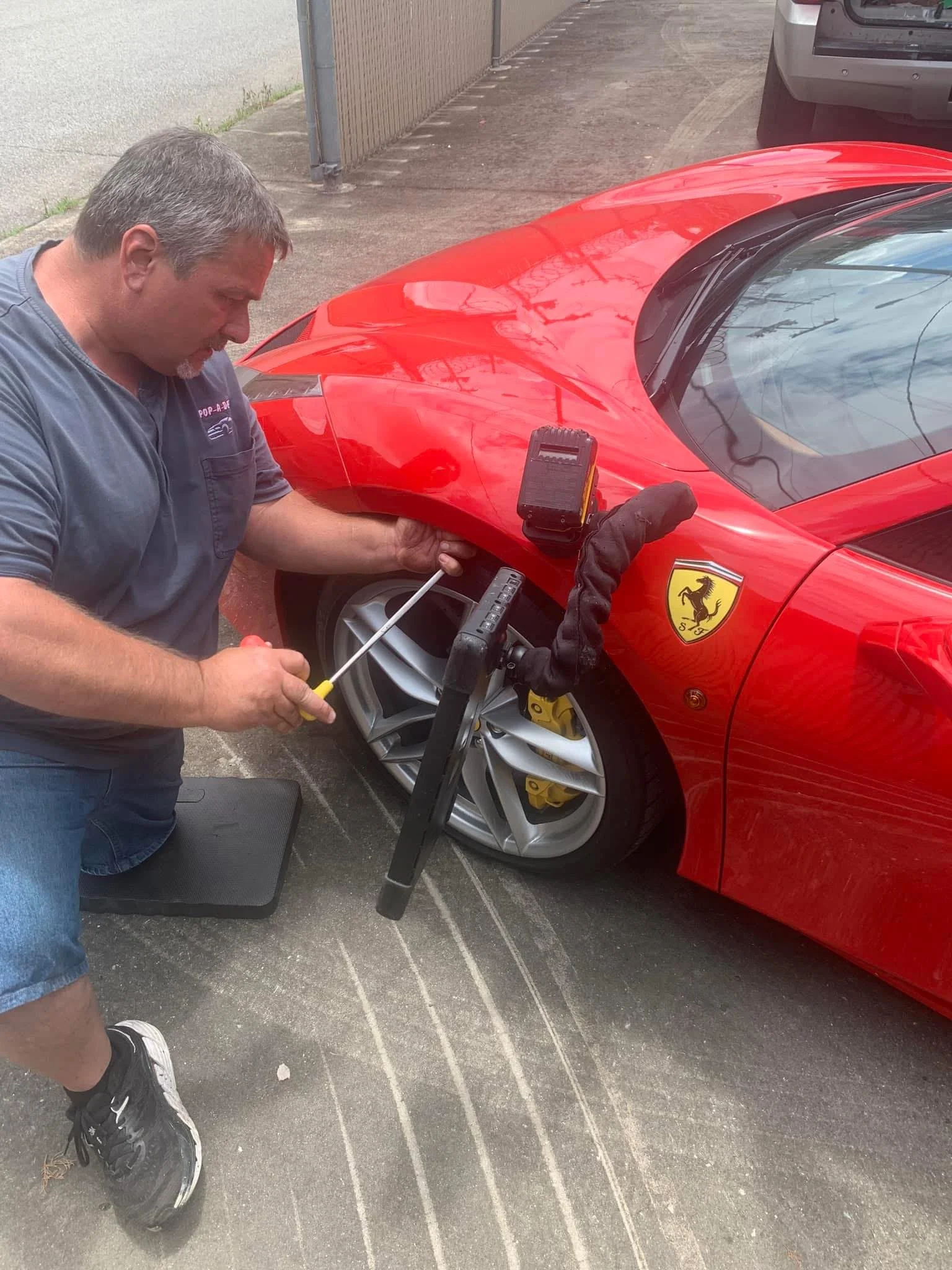 A man in a gray shirt and jeans working on the front wheel of a red Ferrari sports car, parked on a driveway, with a tool and a yellow calibration sticker on the wheel.