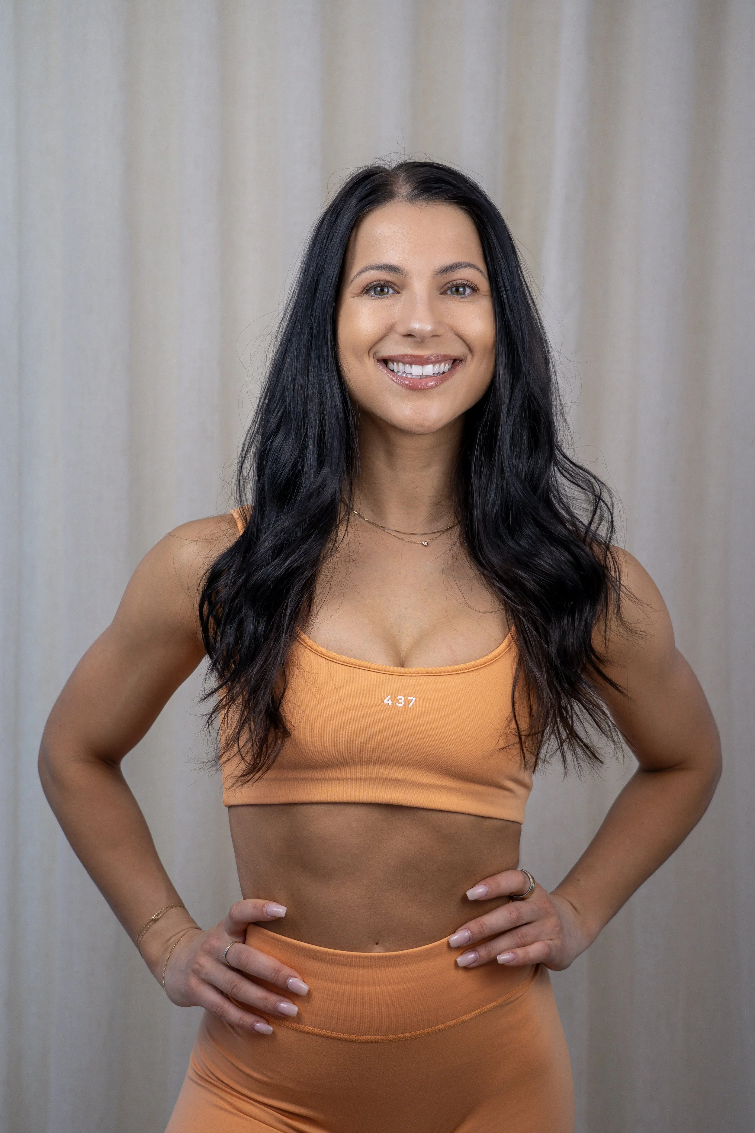 A woman with long dark hair smiling, wearing a beige sports bra with white trim and the number 437, and a matching beige skirt, standing against a plain white background.