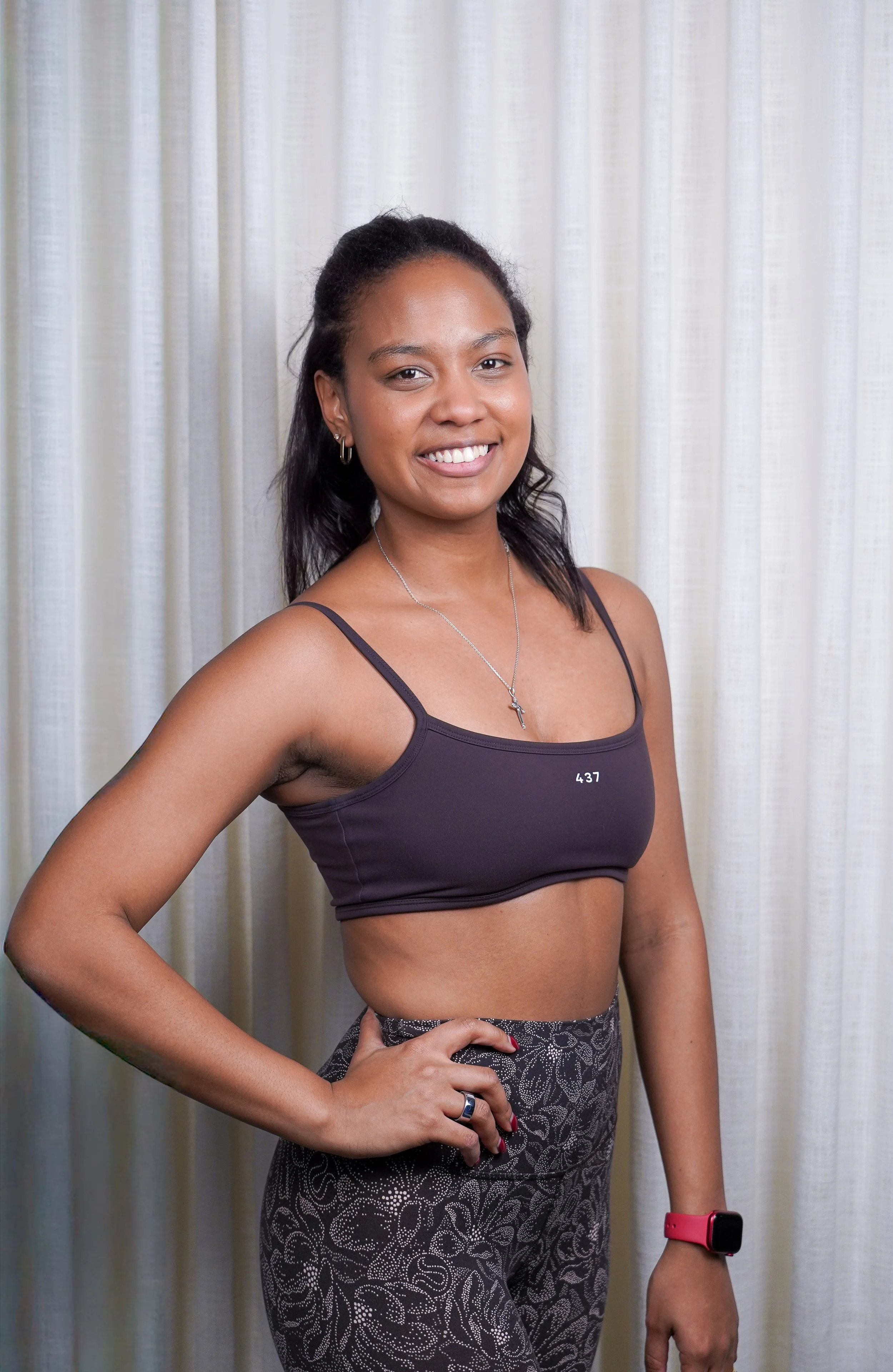 Woman smiling, standing with hands on hips, wearing white sports bra and green athletic shorts, against a plain light background.