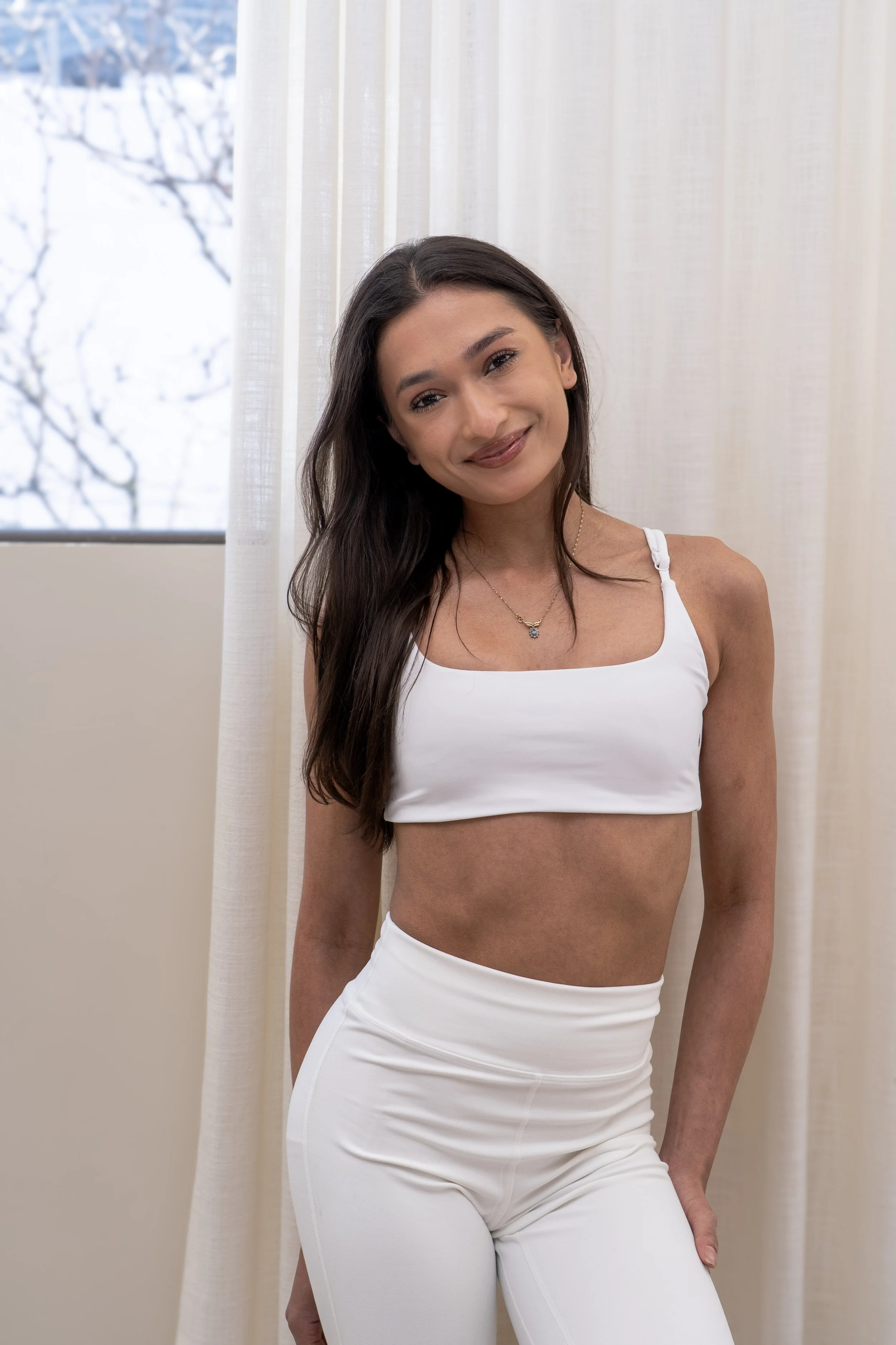 A woman with long dark hair and light skin standing against a plain light wall, wearing teal workout clothes and an Apple Watch.