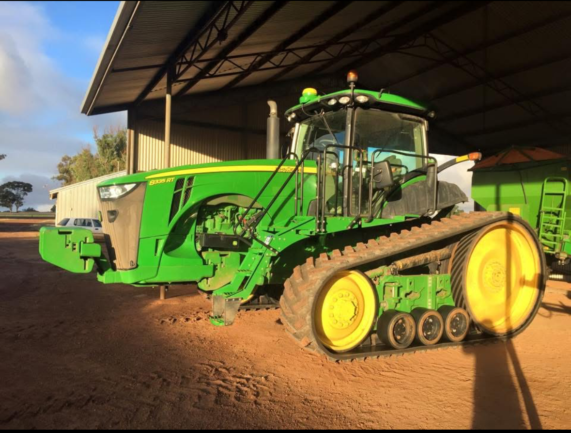 Green agricultural tractor with track wheels parked under a shed with an attached grain cart.