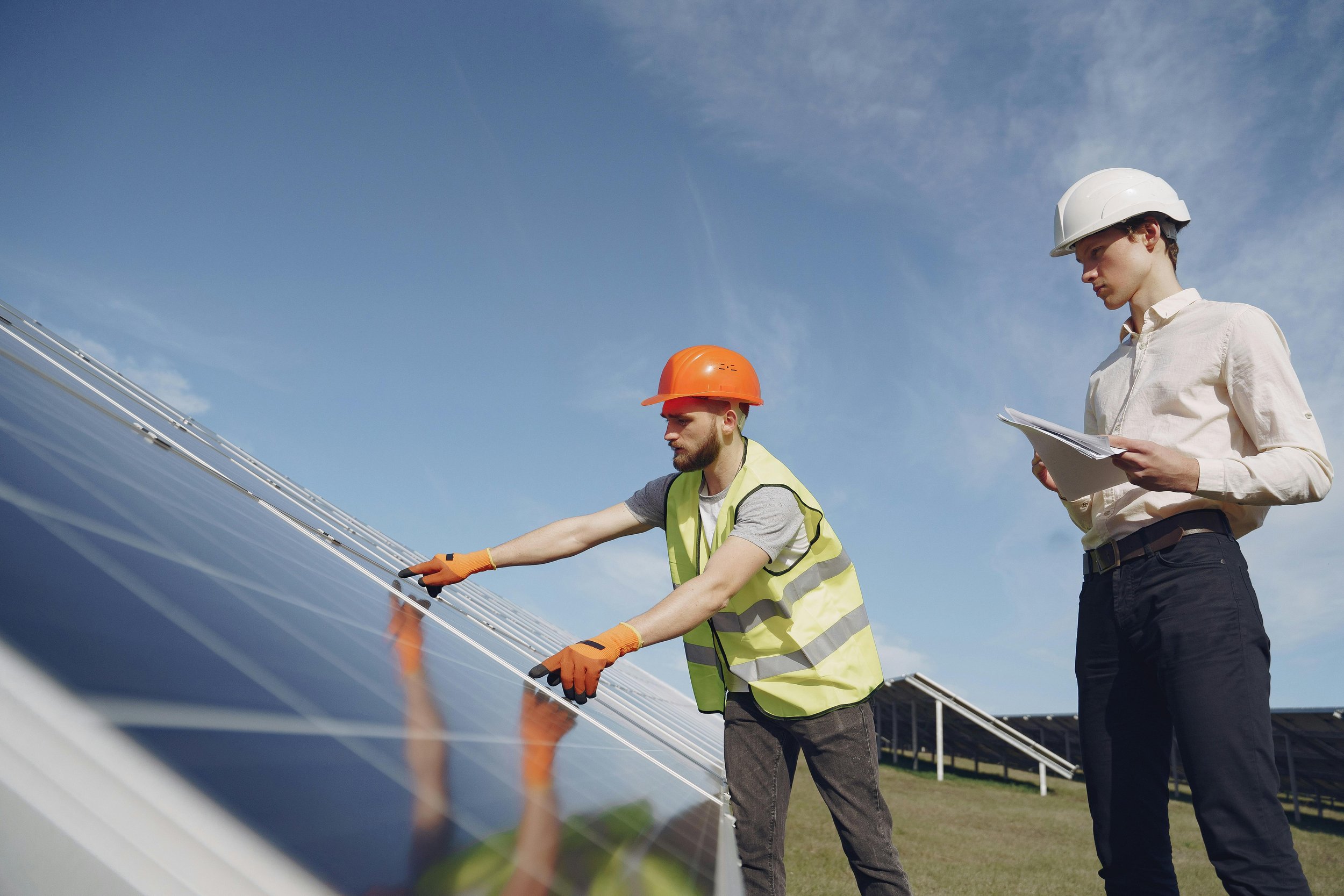 Two electricians installing and inspecting ground-mounted solar panels in a wide grass field, ensuring proper alignment, safety, and efficient system performance.