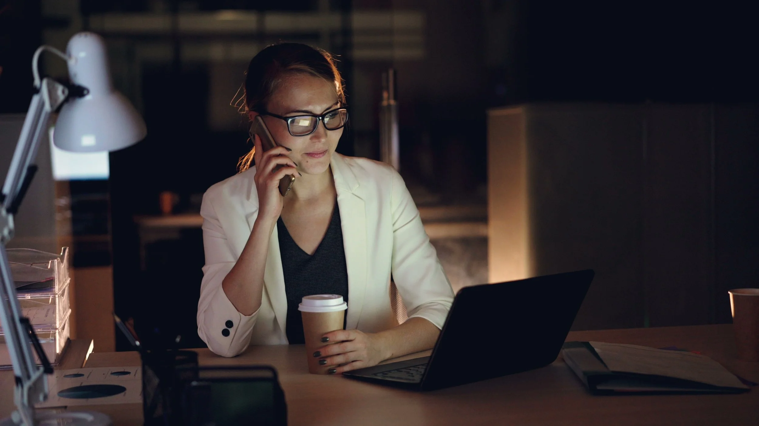 Woman wearing glasses working on a laptop at night while holding a coffee and making a phone call, representing professional focus, reflection, and self-awareness.