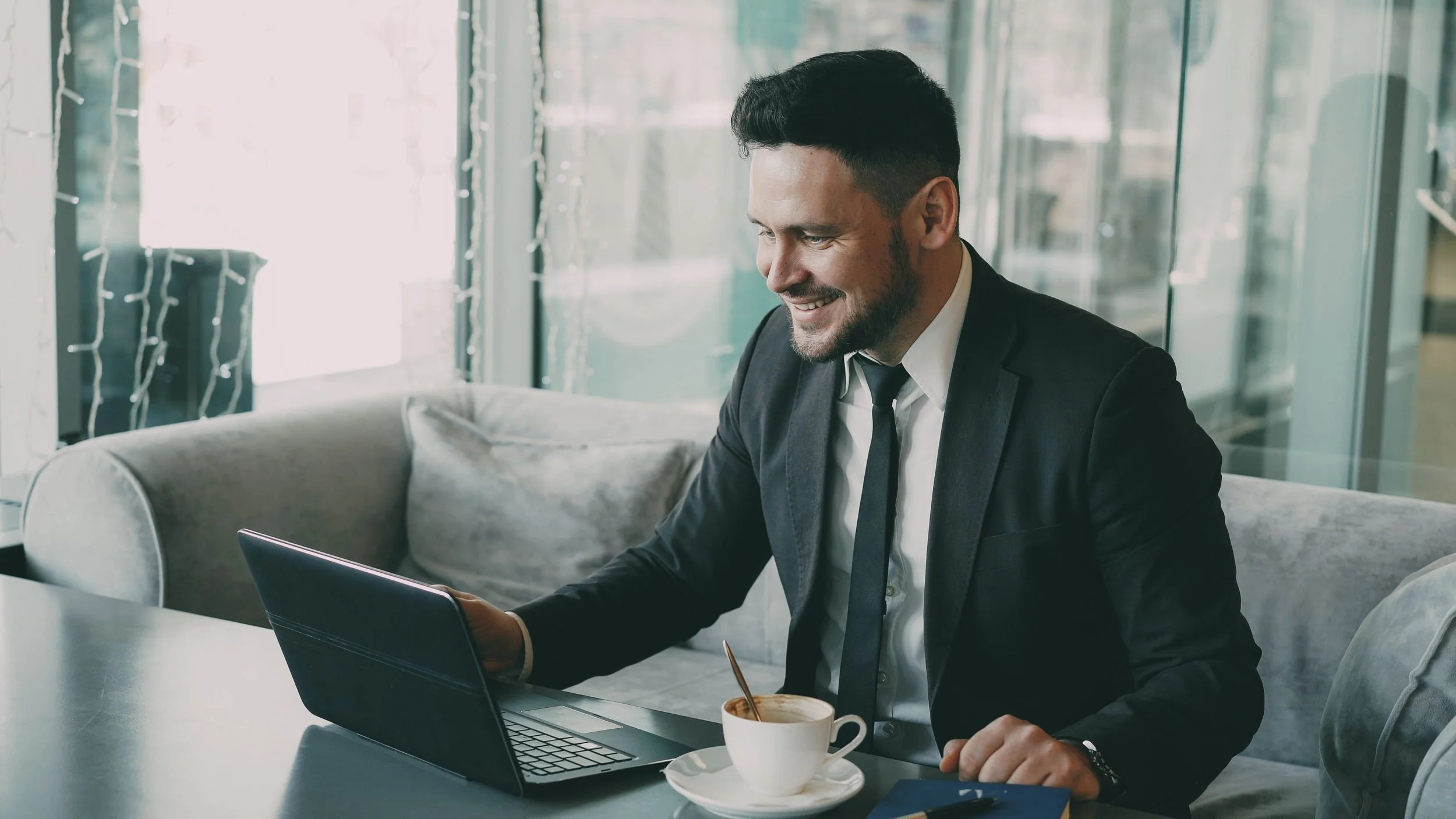 Smiling businessman in a suit working on a laptop while enjoying coffee in a cosy cafe, representing continuous learning, professional growth, and career development.