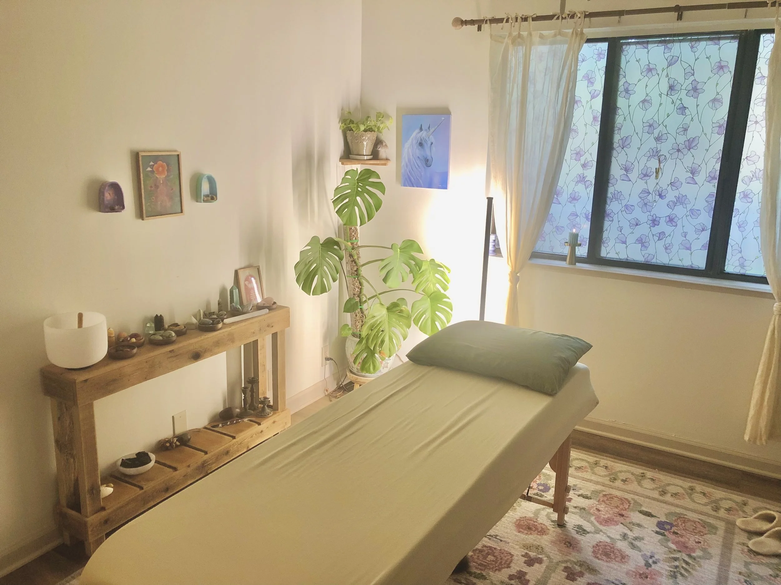 Comfortable massage room with a massage table, a pillow, and a wooden shelf holding crystals, candles, and decor, with a large Monstera plant, a unicorn picture, and floral window curtains.