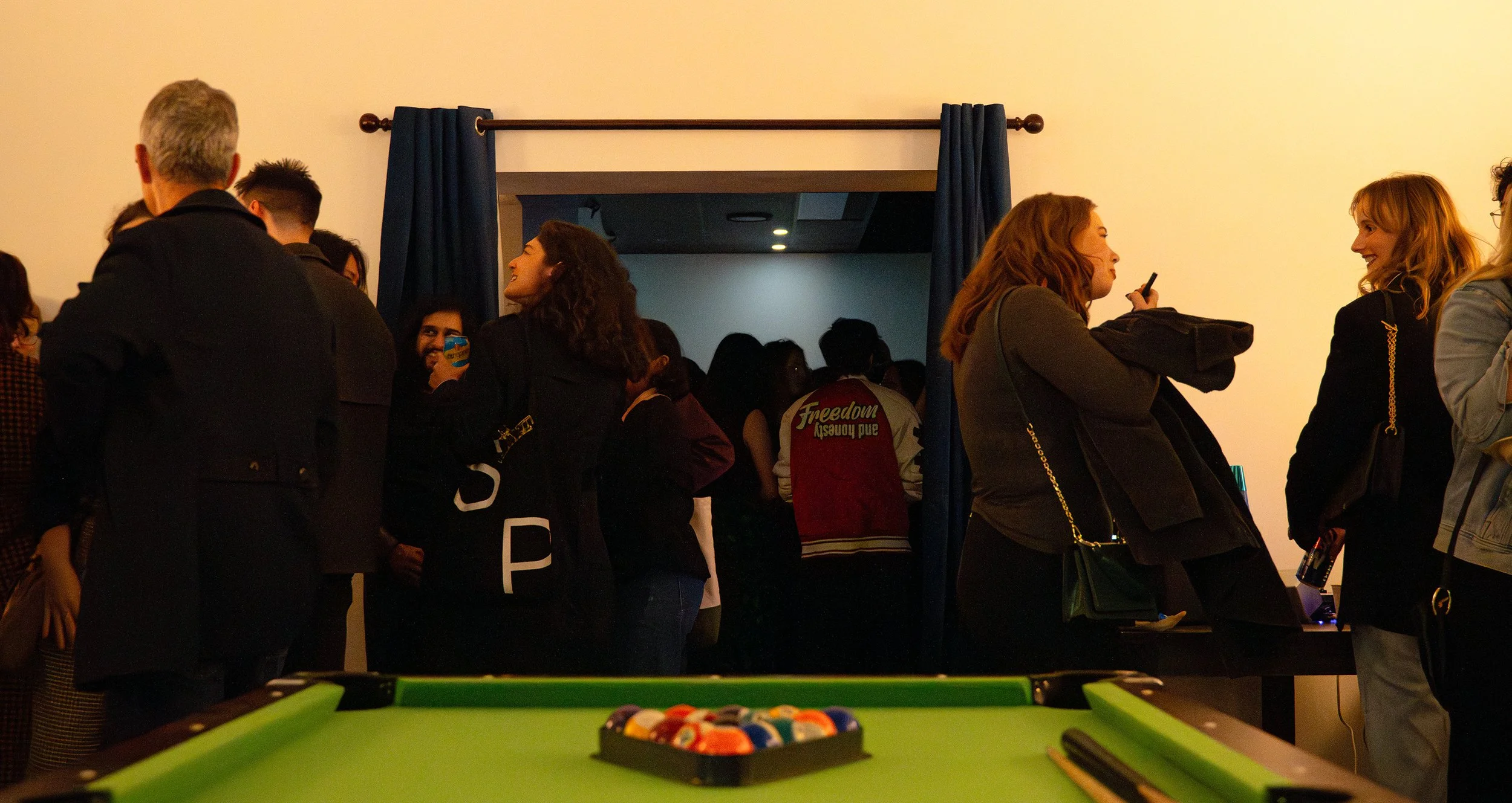 People socializing in a room with a pool table in the foreground, some holding drinks, while others are engaged in conversations.
