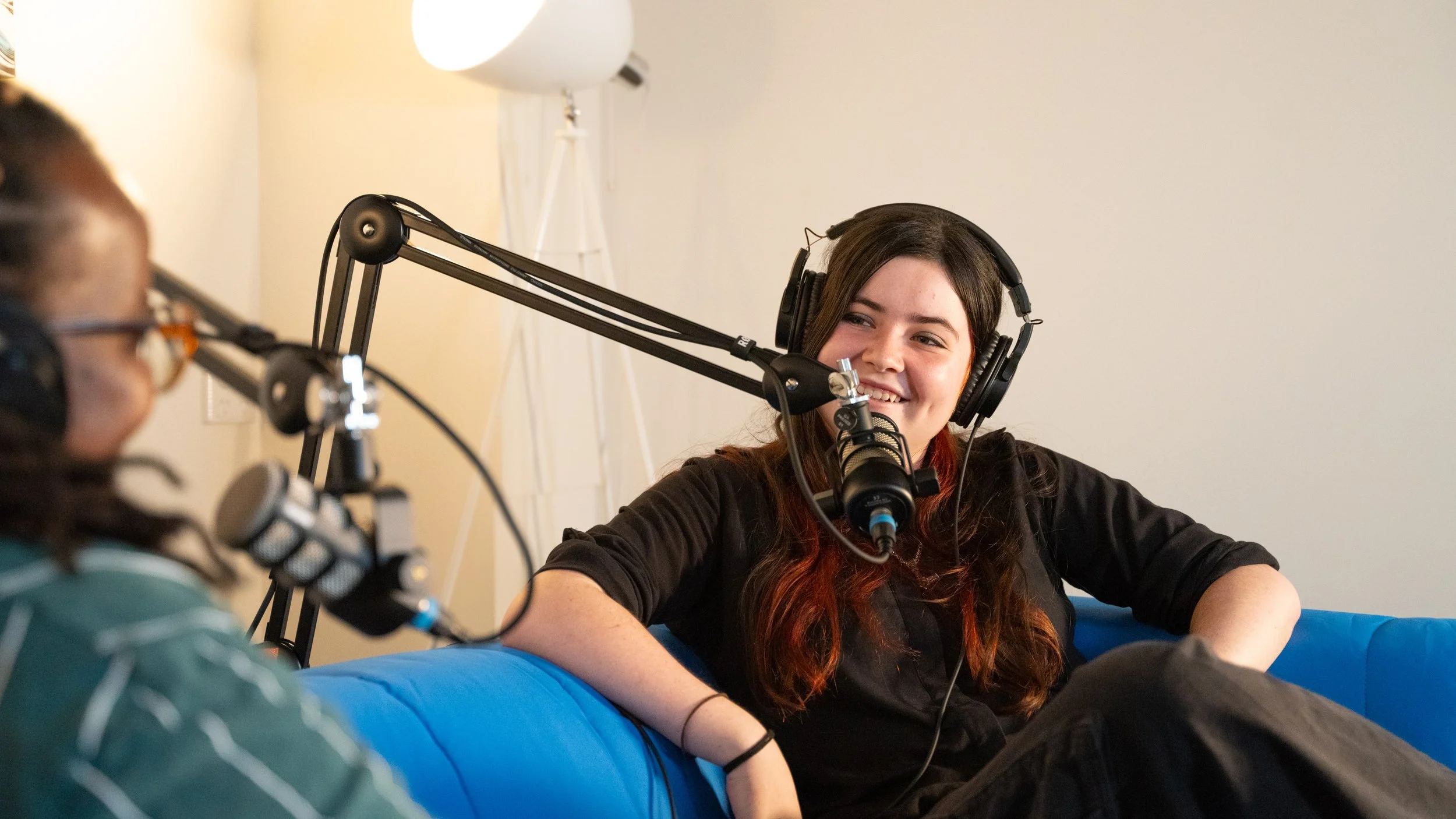 Two women podcasting with microphones and headphones in a room with a white background and a standing lamp.