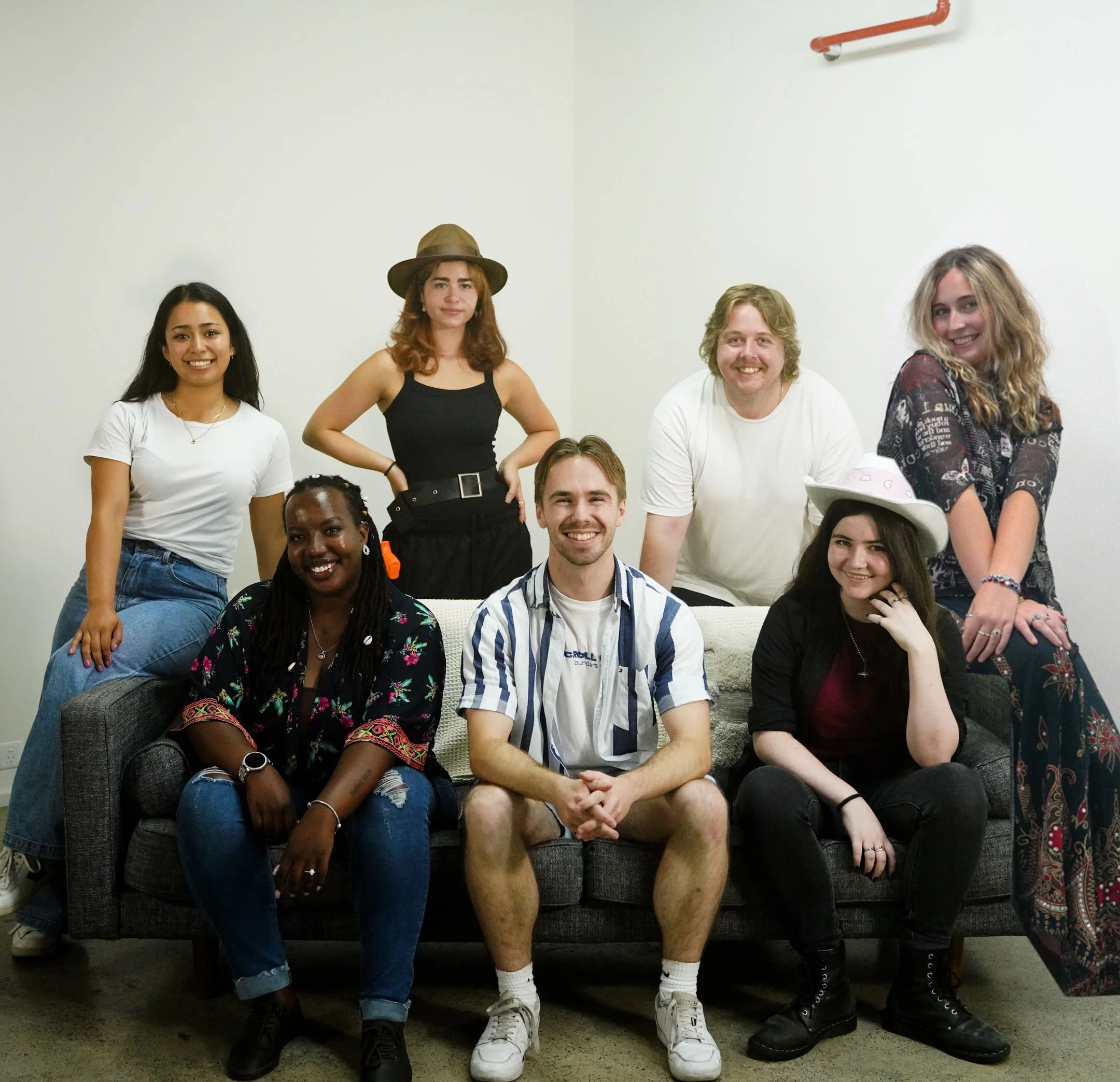 An image of the Scroll Stop team. A diverse group of seven adults, five women and two men, posing together on and around a gray couch against a plain white wall with an orange pipe overhead, smiling at the camera.