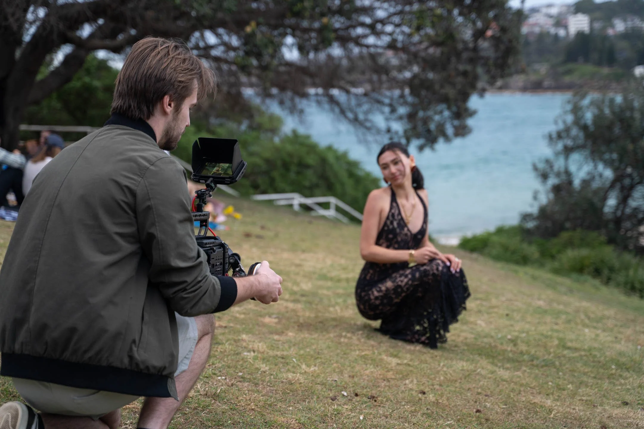 A man operating a camera on a tripod is filming a woman who is kneeling on the grass near the ocean, surrounded by trees and greenery.