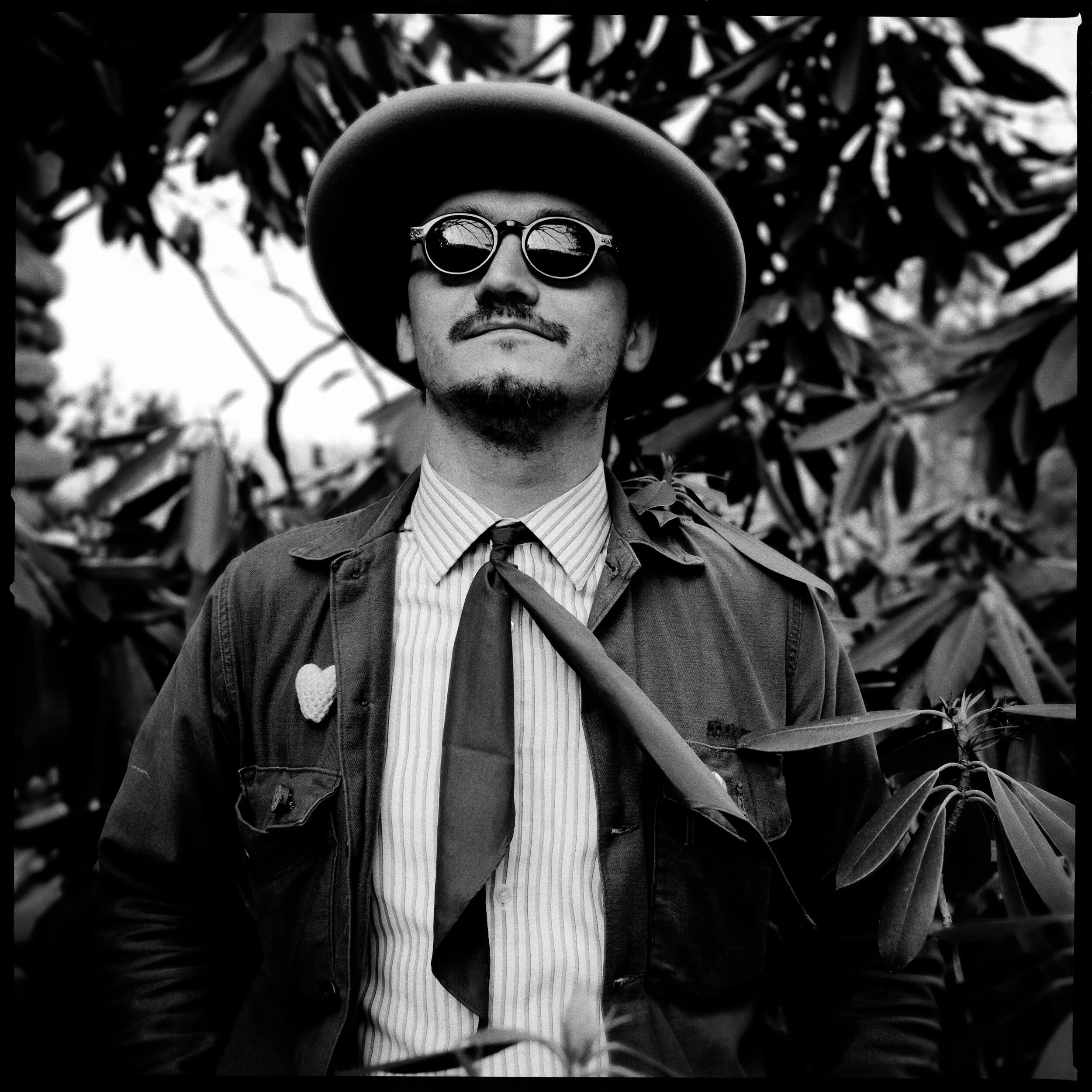 Parker J Pfister Fine Art Black and White Photography of a man standing outside wearing a brimmed hat, sunglasses, and a tie with trees in the background. 