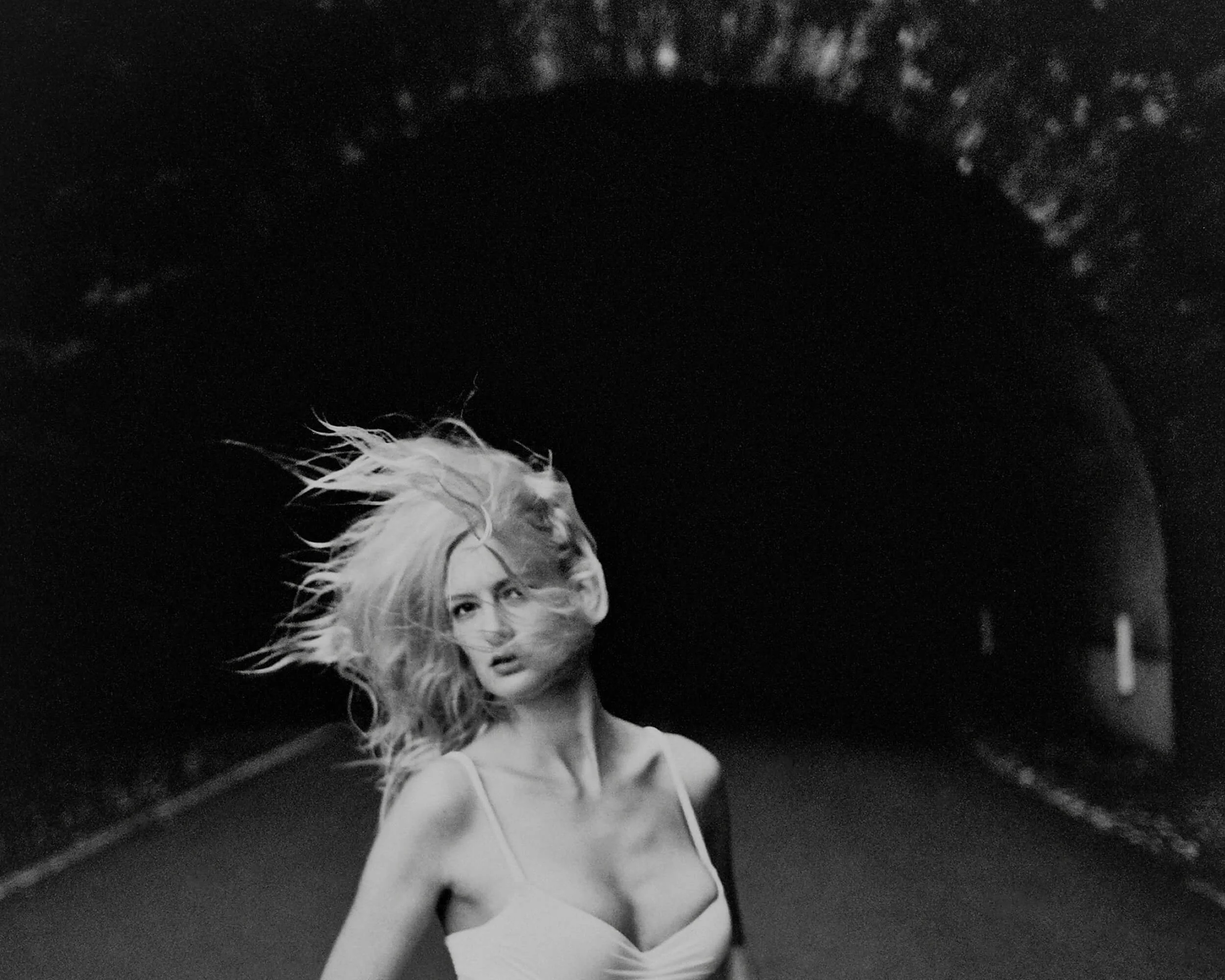 Parker J Pfister Fine Art Black and White Photography of a white woman with her hair blowing in the wind standing in front of a tunnel. 