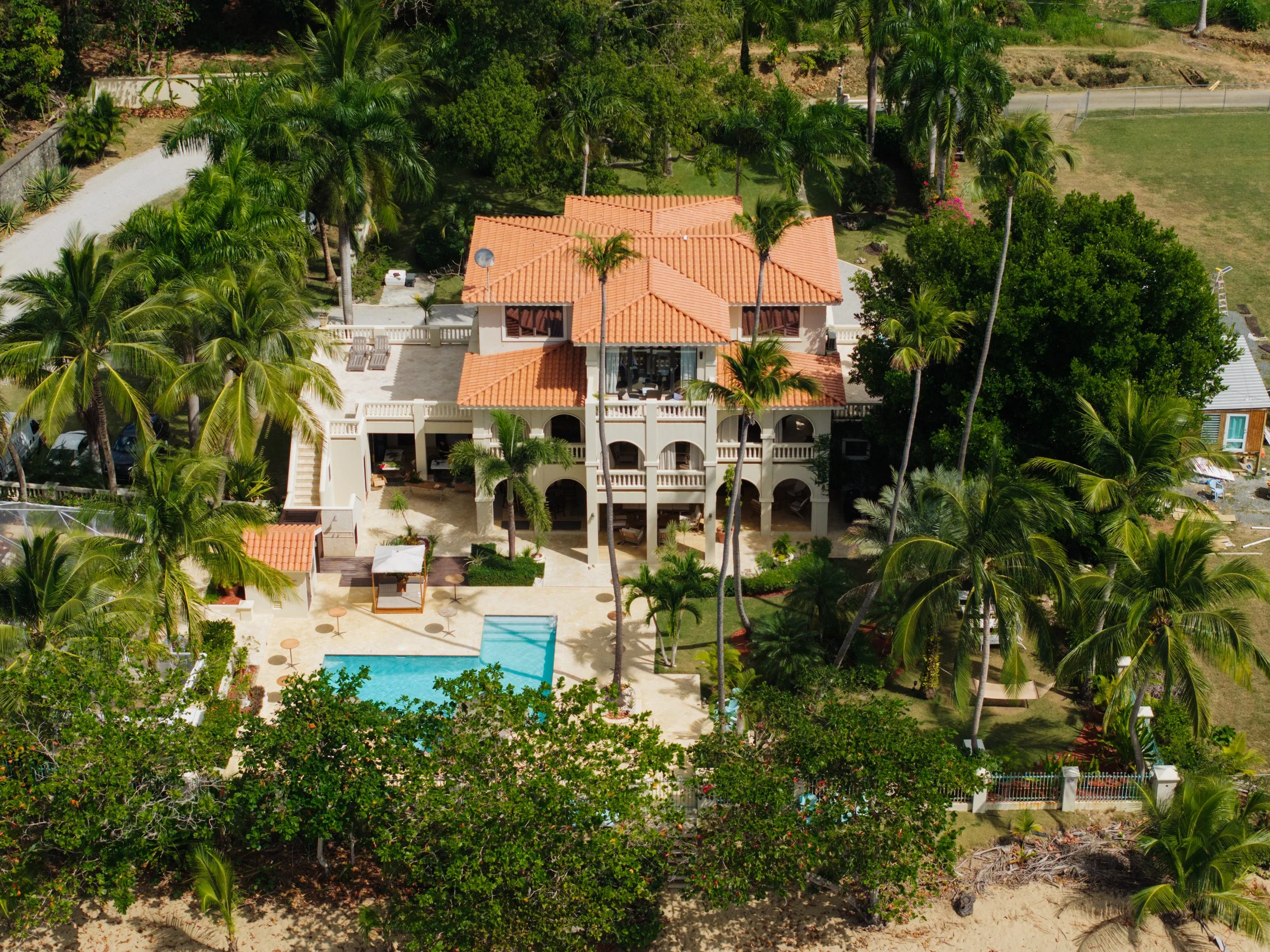 Aerial view of a large tropical house with orange tiled roof, surrounded by palm trees and lush greenery, with a swimming pool and outdoor seating area in the backyard.