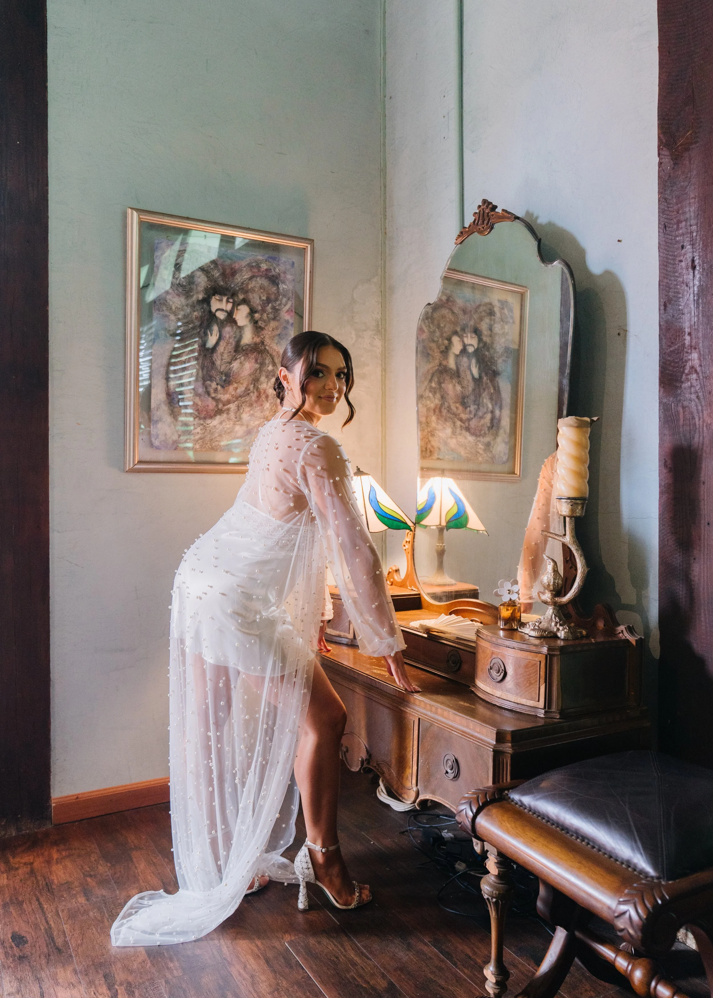 A woman in a sheer, beaded white gown and high heels is leaning on an antique wooden dresser in a vintage-style room, with artwork and a decorative lamp in the background.