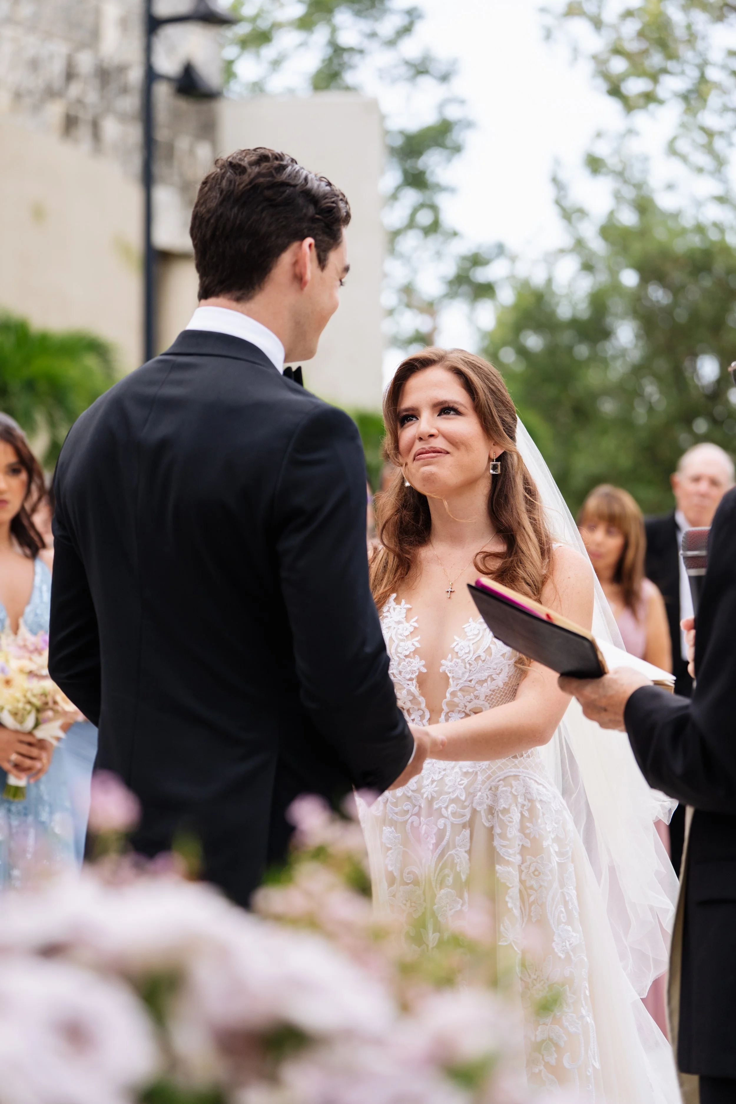 A bride and groom exchanging vows during an outdoor wedding ceremony, with the officiant holding a book, and bridesmaids and guests watching in the background.