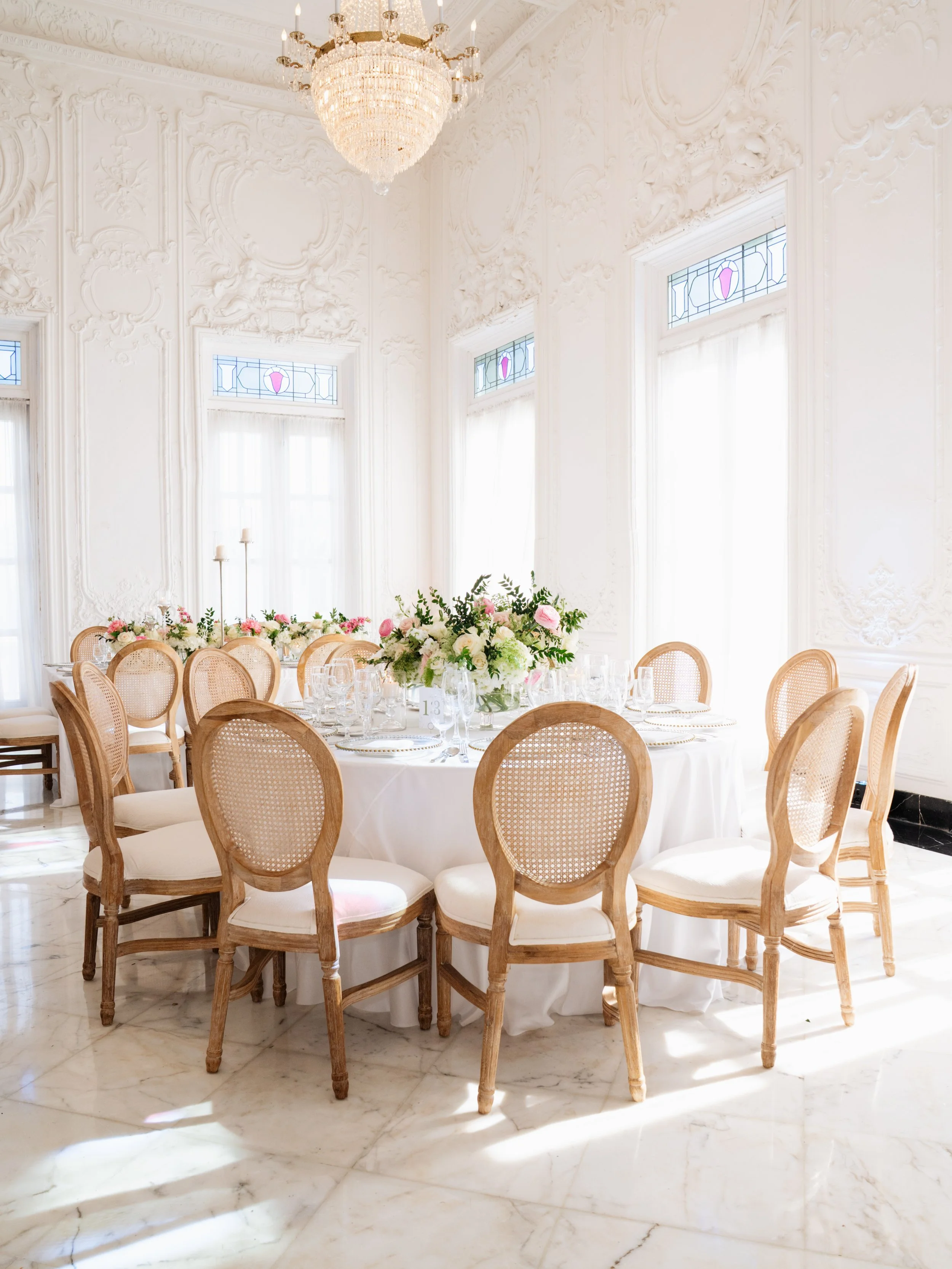 Elegant round dining table set for a formal event with white tablecloth, floral centerpiece, and multiple wine glasses in a bright, ornate room with large windows, chandelier, and white walls with intricate molding.