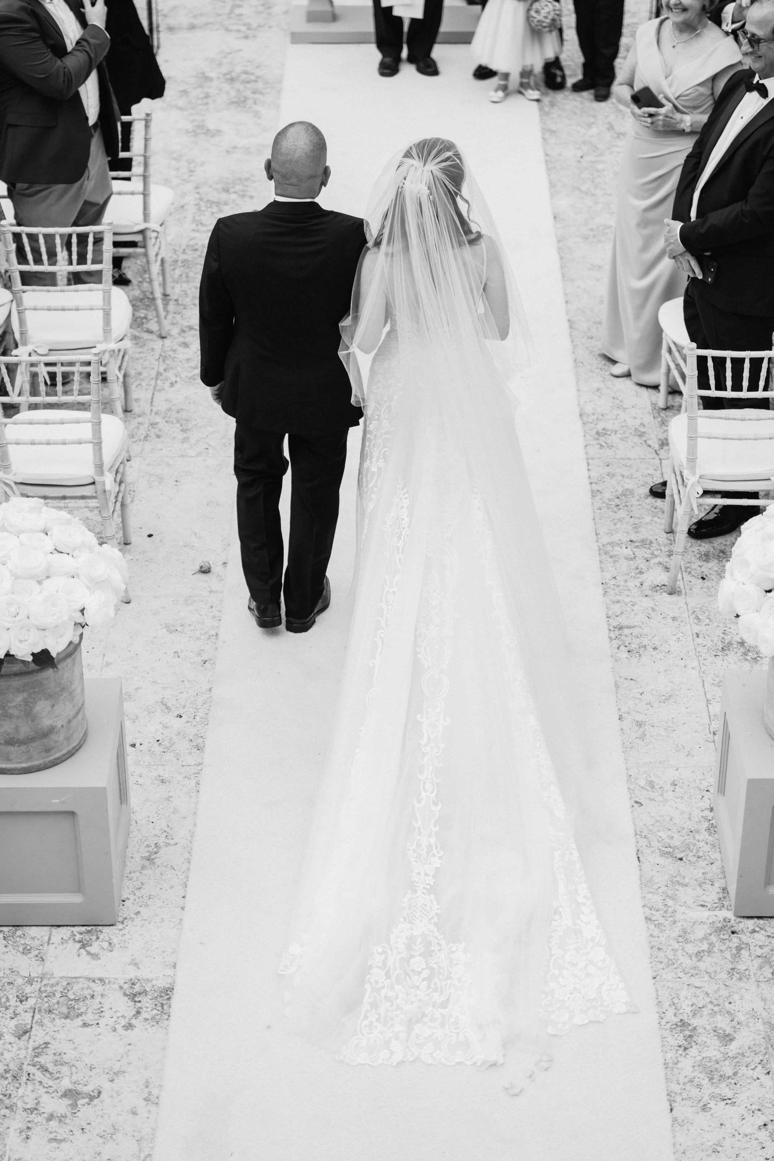 Black and white photograph of a bride and groom walking down the aisle at their wedding ceremony, with guests standing on either side of the aisle.
