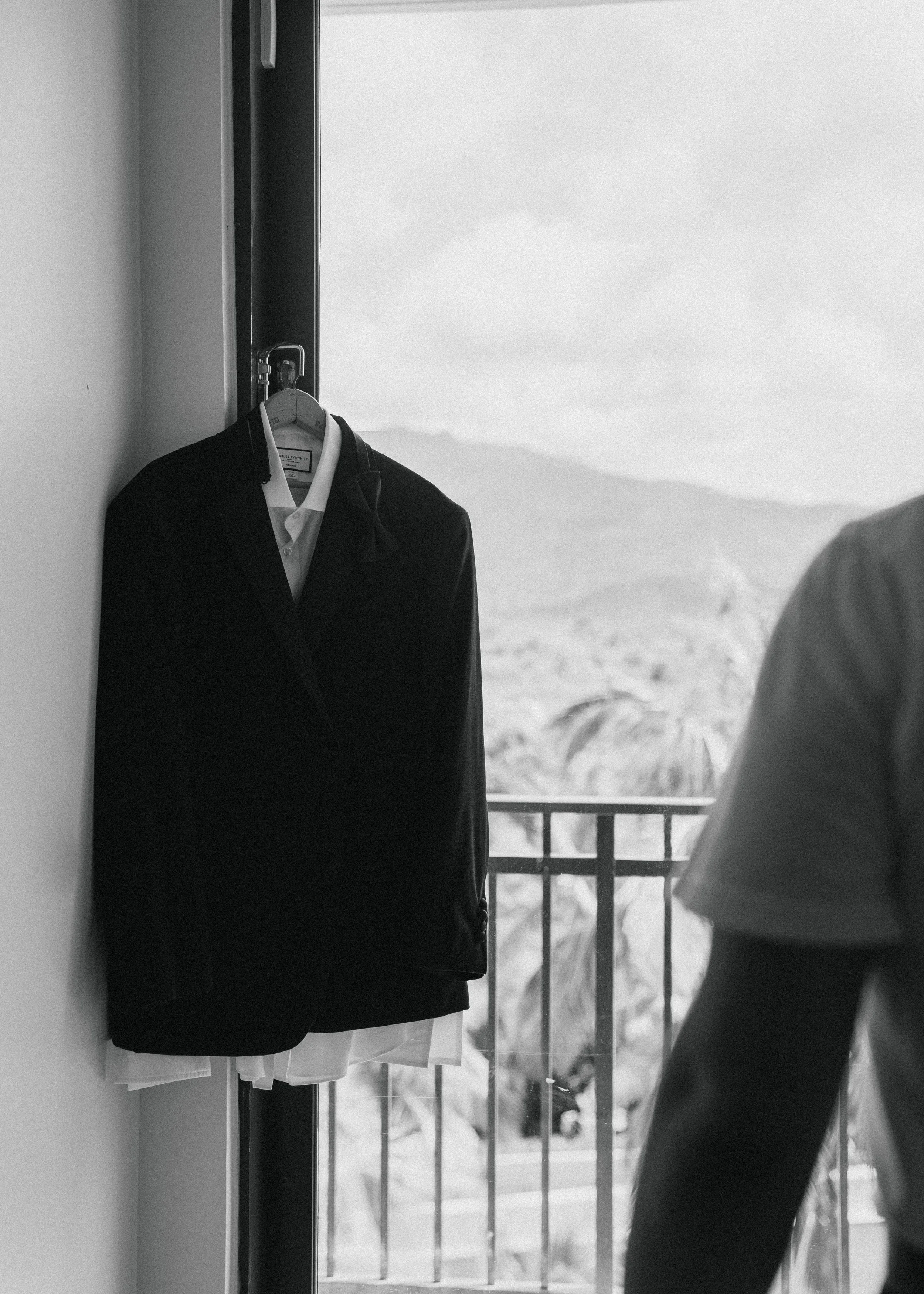 A black suit jacket hanging on a hanger attached to a door frame with a view of a balcony and mountain landscape in the background.