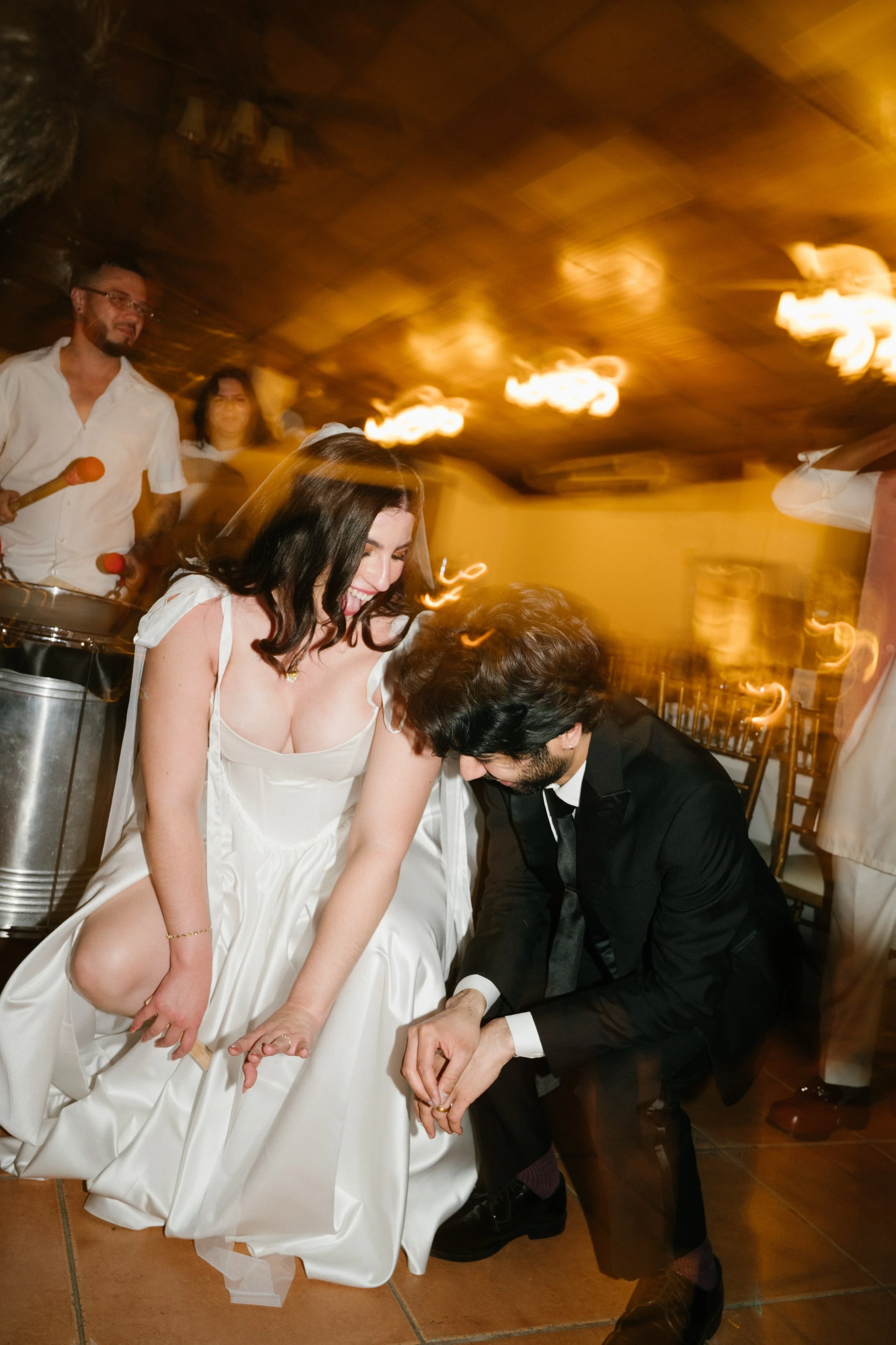 Couple at wedding dance, woman in wedding dress smiling, man in tuxedo, people in background, lively atmosphere with warm lighting.