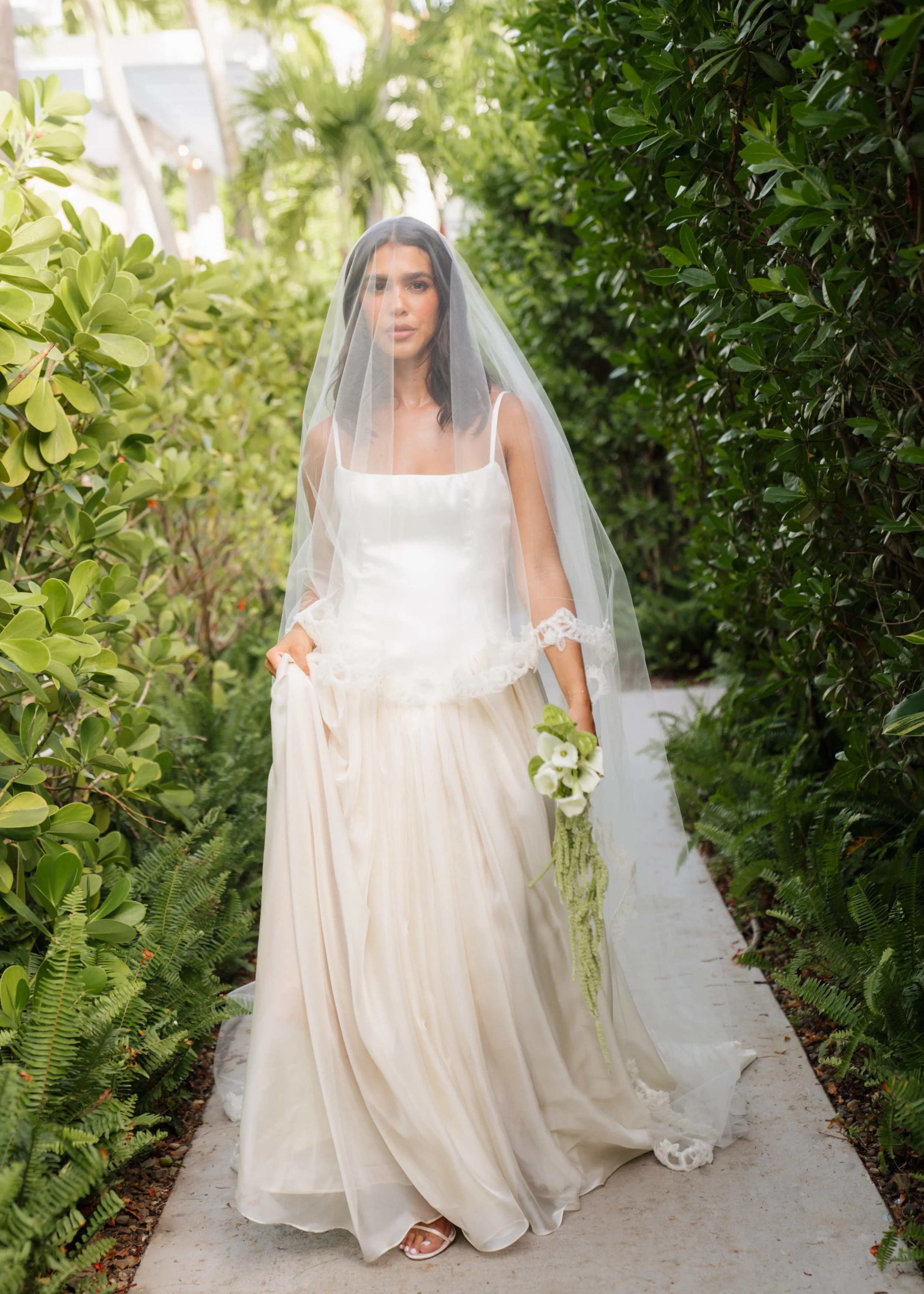 A woman in a wedding dress and veil holding a bouquet walks down a garden path surrounded by green foliage.