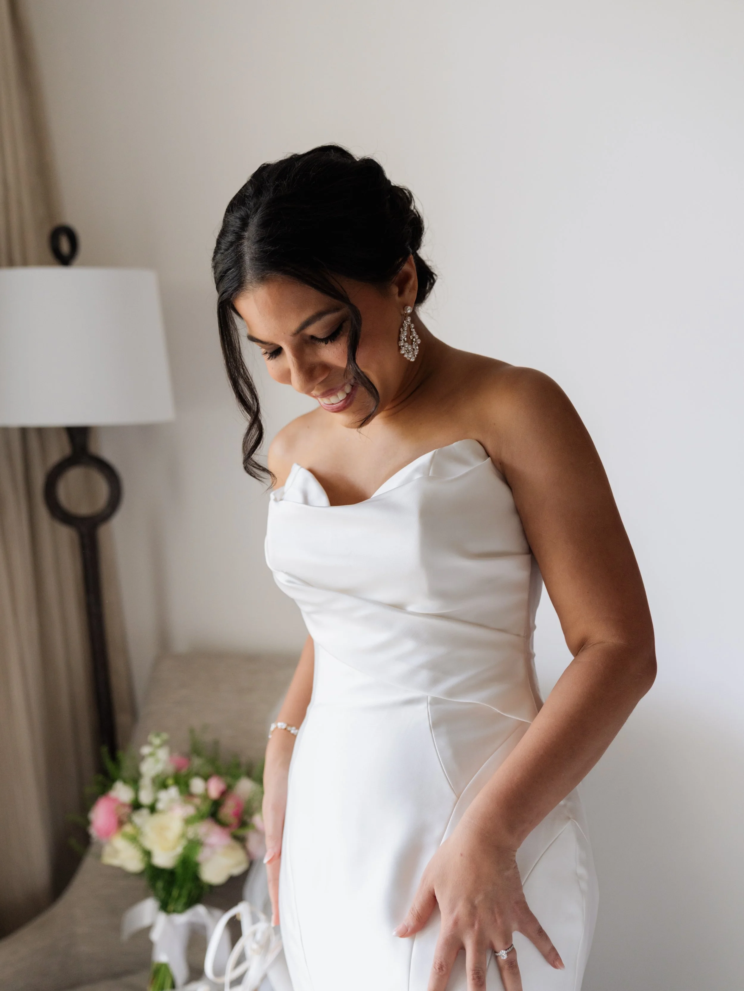 Bride in a white wedding dress smiling and looking down, standing inside a room with a flower bouquet on a chair behind her.