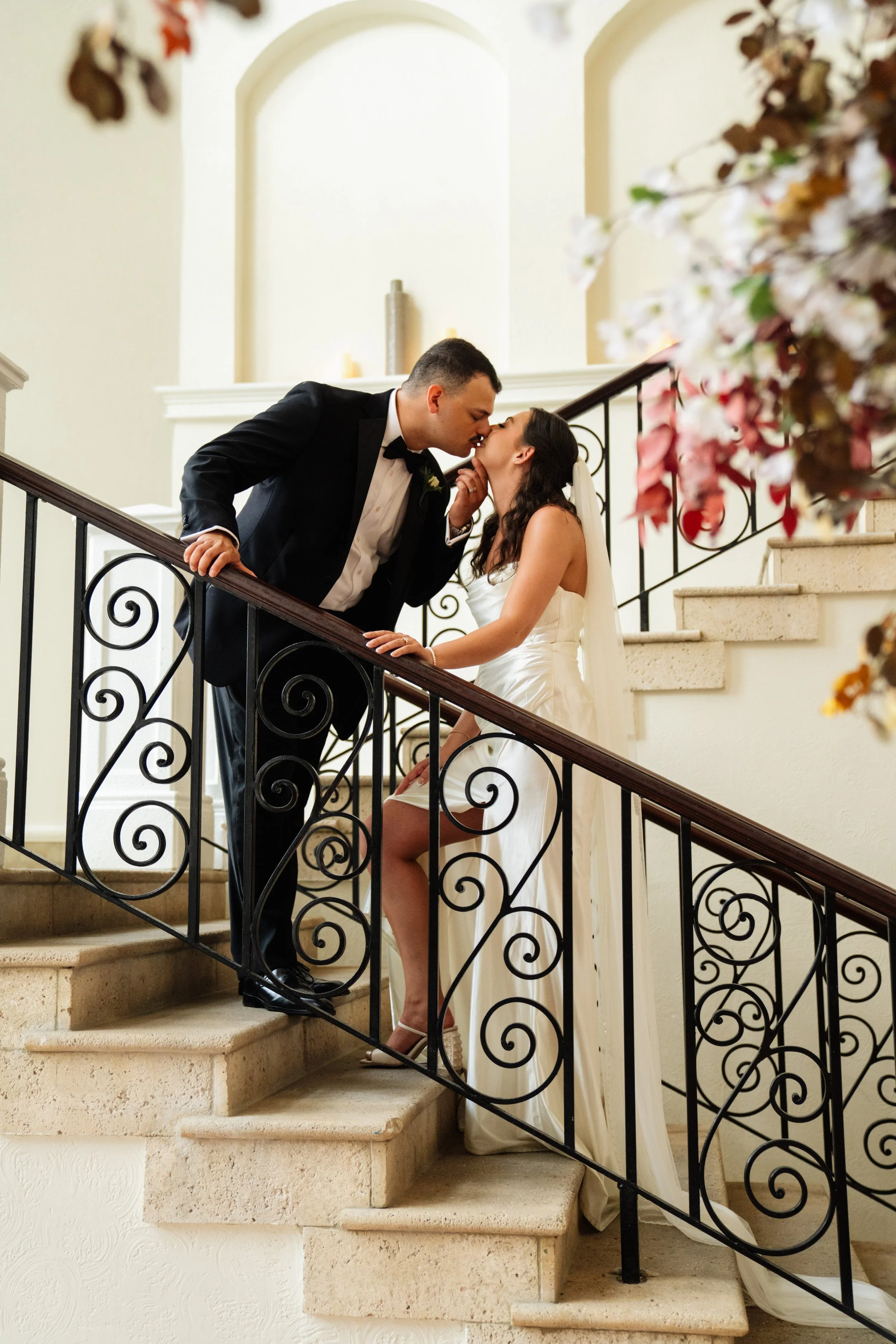 A newlywed couple kissing on a staircase, with the groom leaning down toward the bride, who is sitting on the stairs, in an indoor setting with decorative railing and floral arrangements.