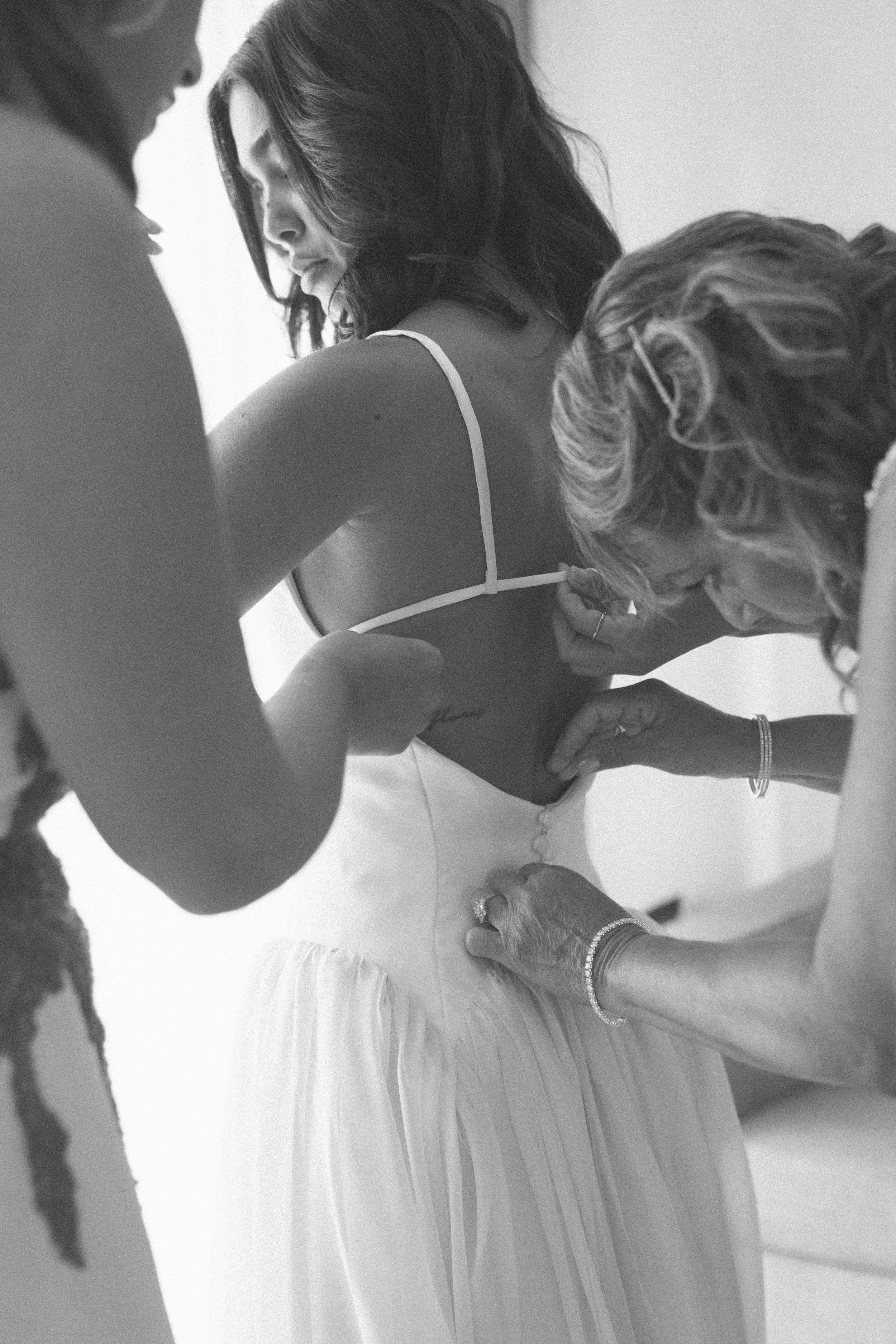 A woman in a wedding dress having her back tattooed by a tattoo artist with the help of two other women.