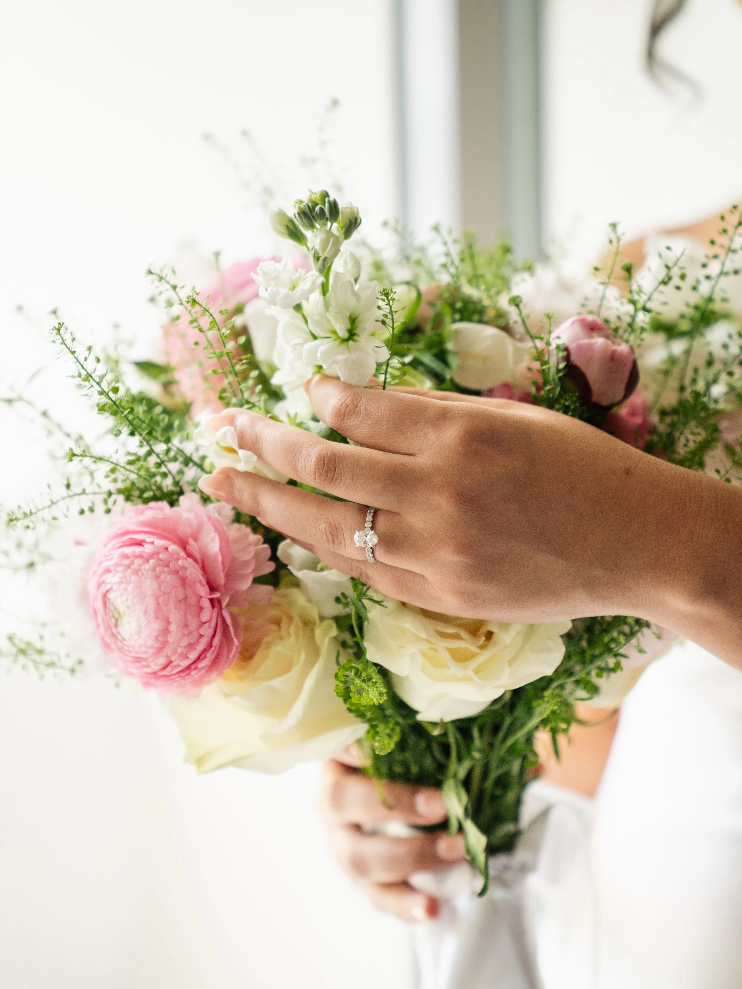 A hand wearing a diamond engagement ring holding a bouquet of white, pink, and green flowers.