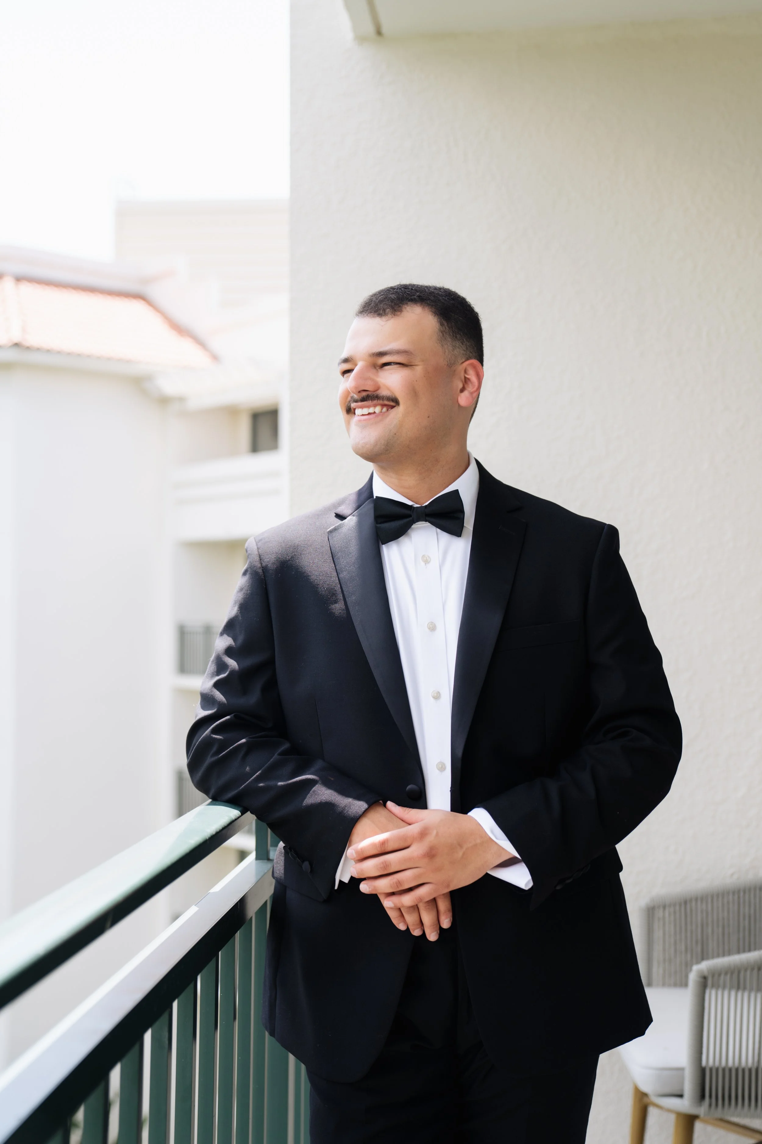 A man in a tuxedo with a black bow tie and white shirt standing on a balcony, smiling and looking to the side.