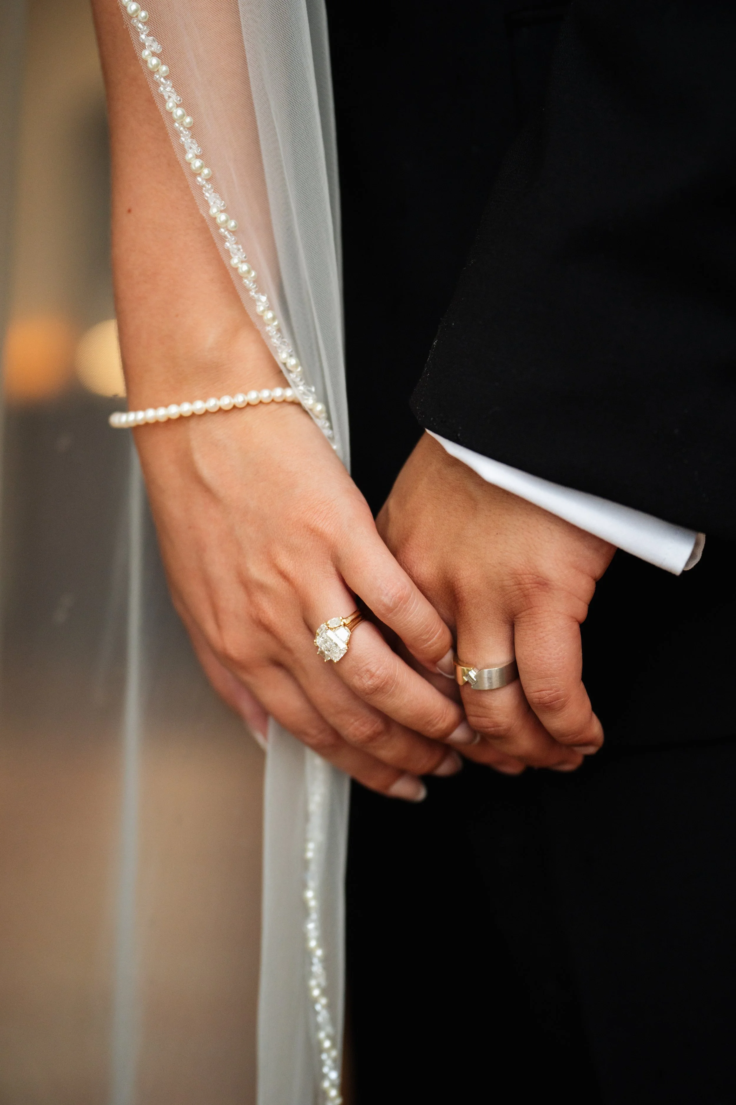 Close-up of a bride and groom holding hands, showing wedding rings; the bride's hand has a large gemstone ring, and she wears a pearl bracelet and a veil with pearl trim, while the groom's hand has a plain metallic band; both are dressed in formal wedding attire.