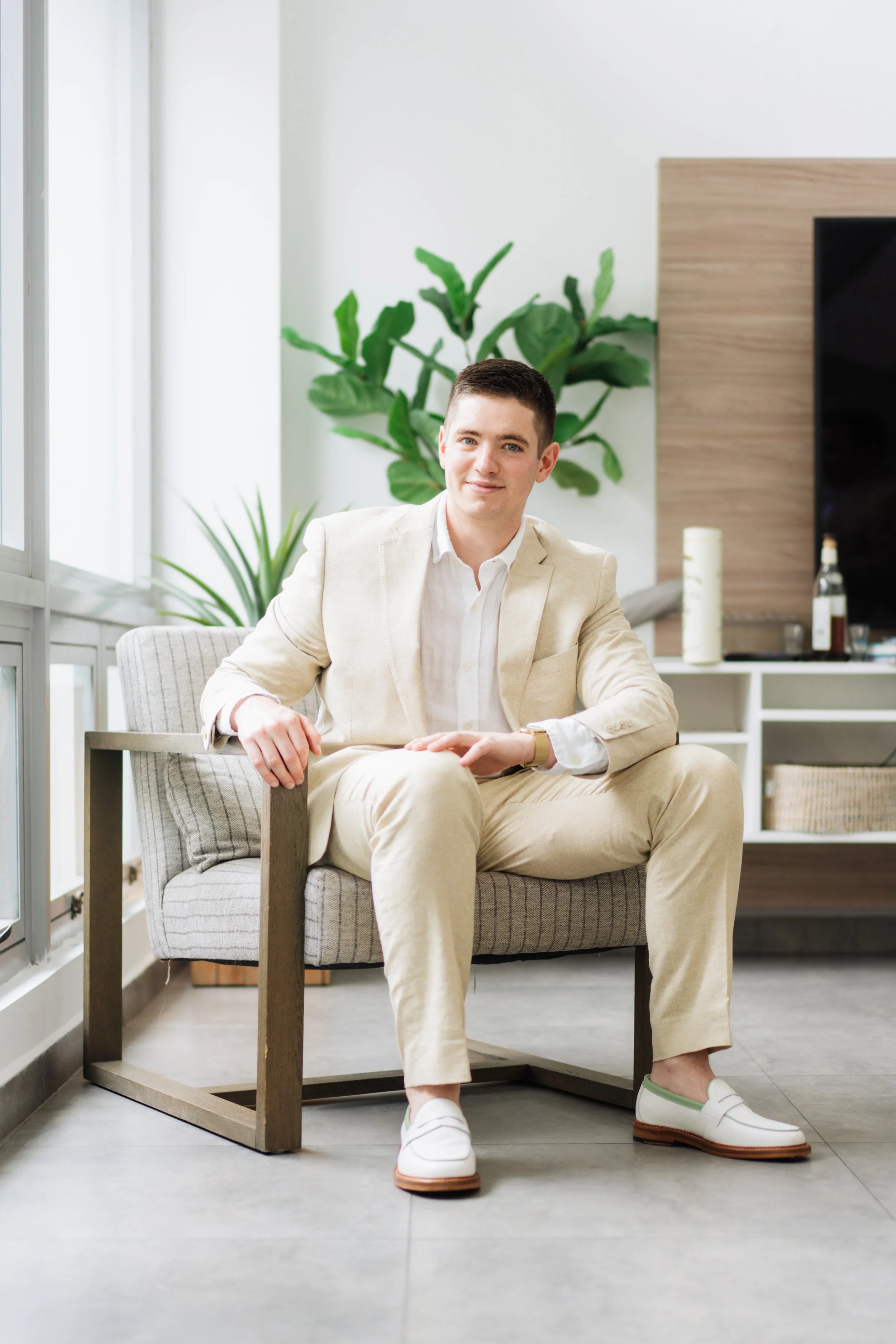 A man in a cream suit with a white shirt sitting on a modern gray chair in a bright indoor space, with large green plants and a wooden shelf in the background.