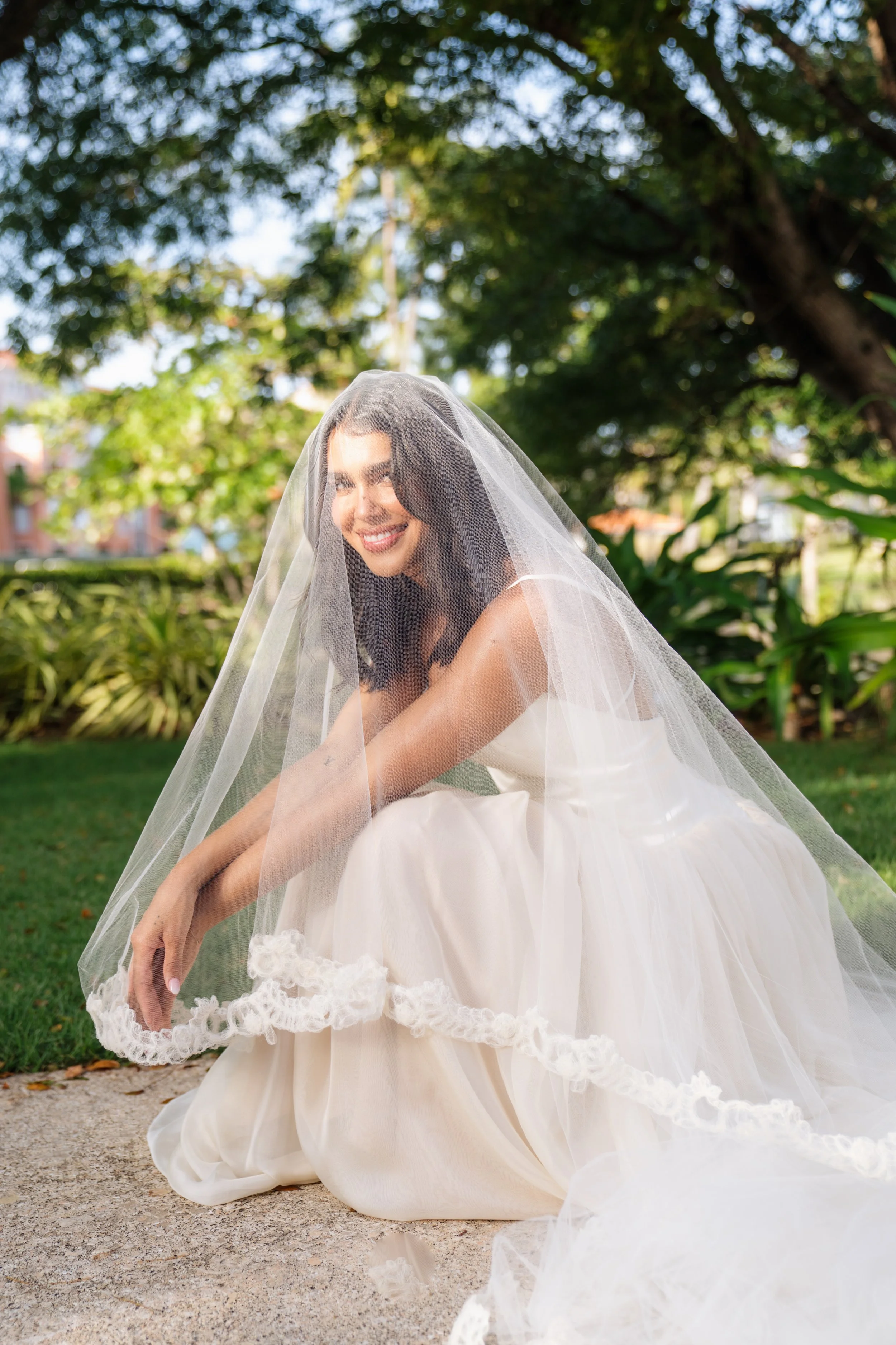A woman in a wedding dress with a veil, sitting on the ground outdoors and smiling at the camera.