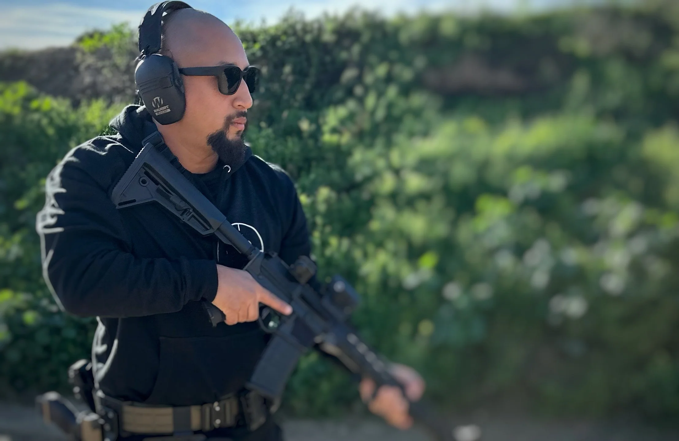 A man wearing sunglasses, protective headphones, and a black hoodie holding a rifle outdoors with greenery in the background.