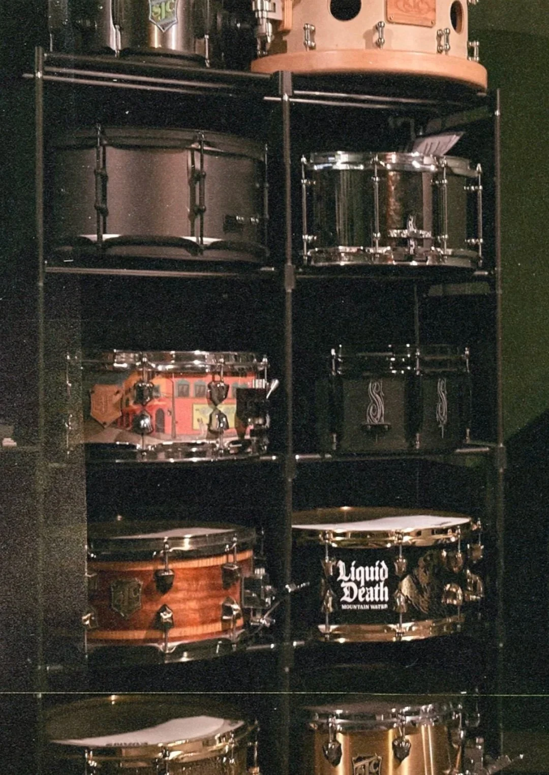 A black shelving unit displaying drums and percussion instruments, including a black snare drum, a brown snare drum with a logo, and a black drum with a liquid death sticker, along with some cymbals and hardware.