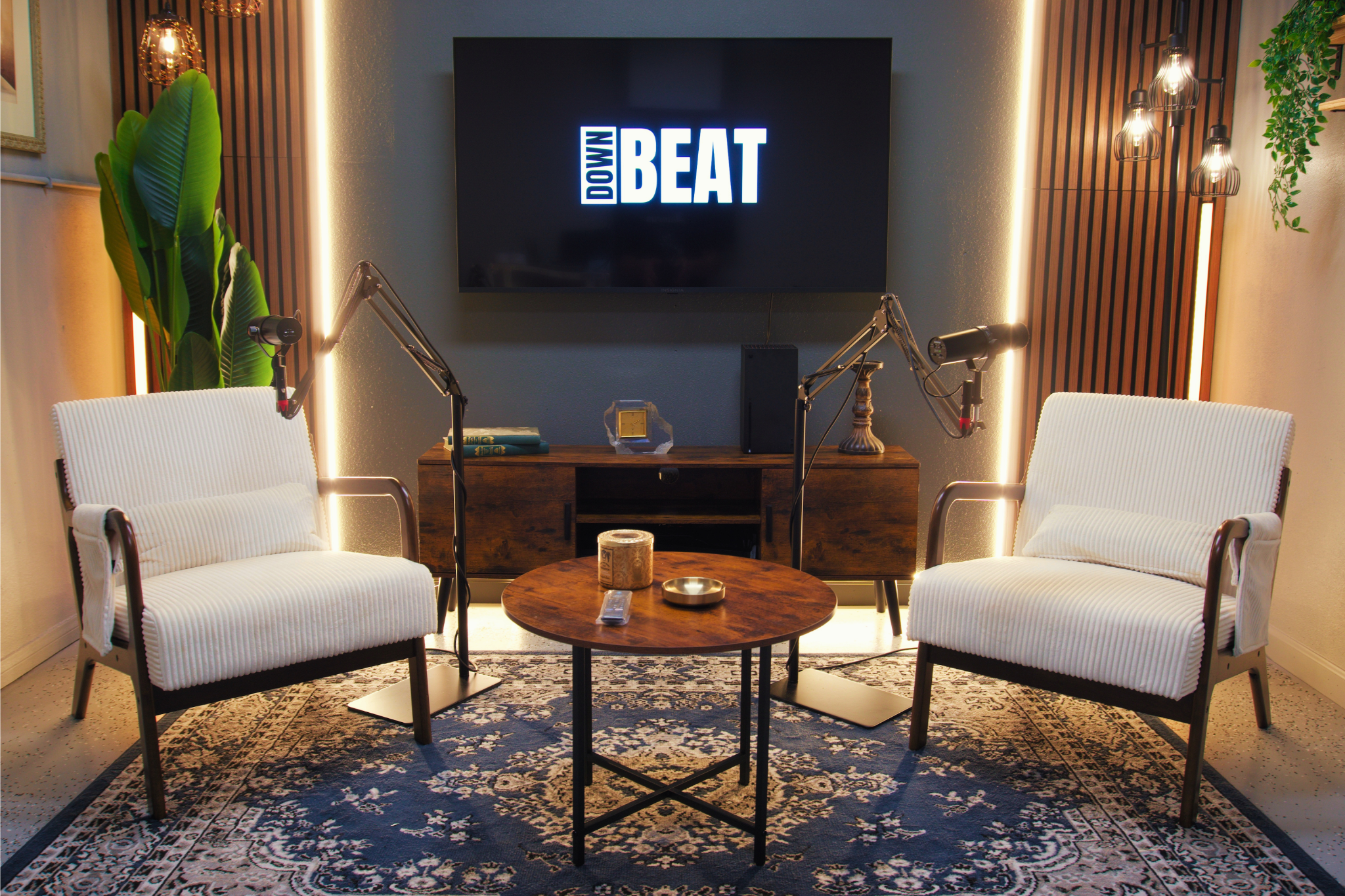 A cozy podcast recording room with two white armchairs facing each other, a round wooden coffee table in the center, a large flat screen TV on the wall displaying DOWI BEAT, two microphones on adjustable stands, a vintage-style table lamp, a potted plant, and decorative lighting along the walls.