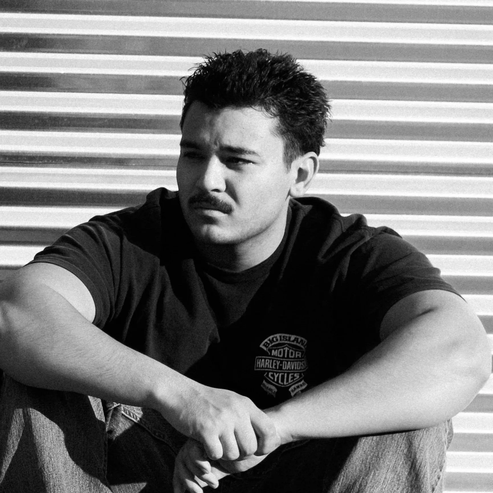 Black and white photo of a man with a mustache and styled hair, wearing a Harley-Davidson t-shirt, sitting against a corrugated metal background, with his arm resting on his knee.