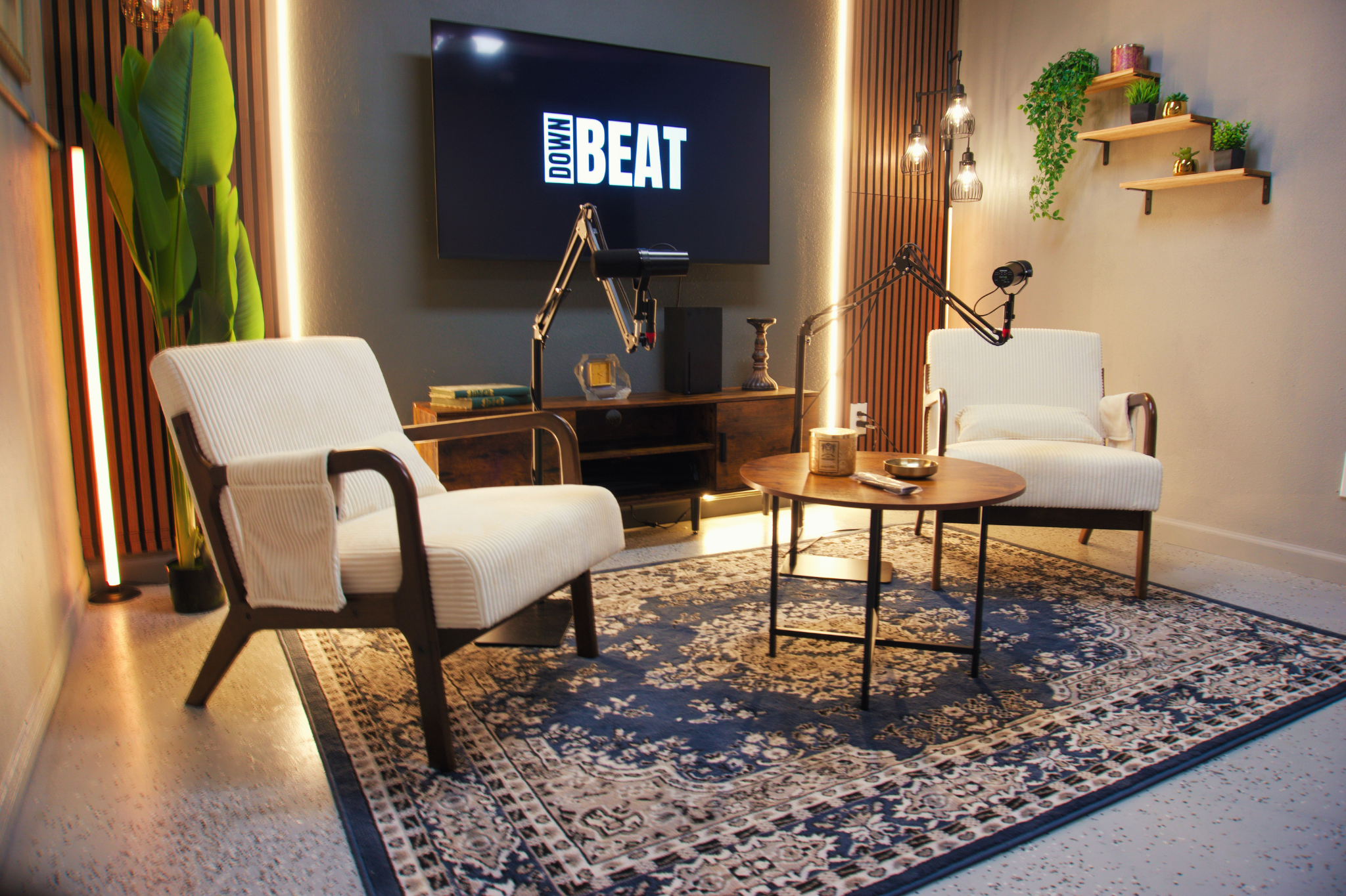 A cozy living room set up for recording or streaming, with two white armchairs, a small round wooden table, a large flat-screen TV displaying the words 'Dolly Beat,' and decorative shelves with plants on the wall. The room has warm lighting, a patterned rug, and modern decor including hanging light fixtures and curtain panels.