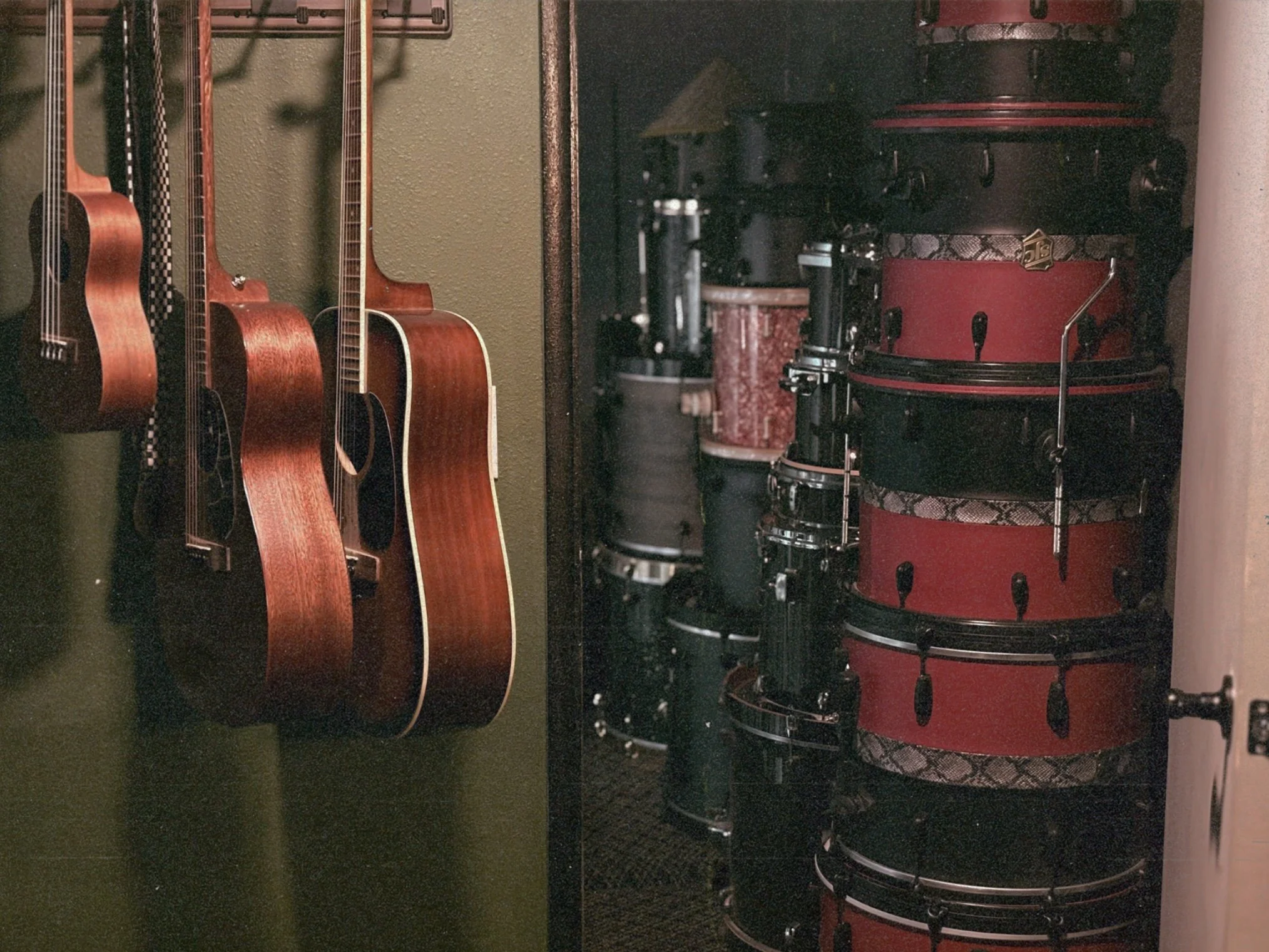 Several acoustic guitars hanging on a green wall next to a red drum set with two drums and cymbals.