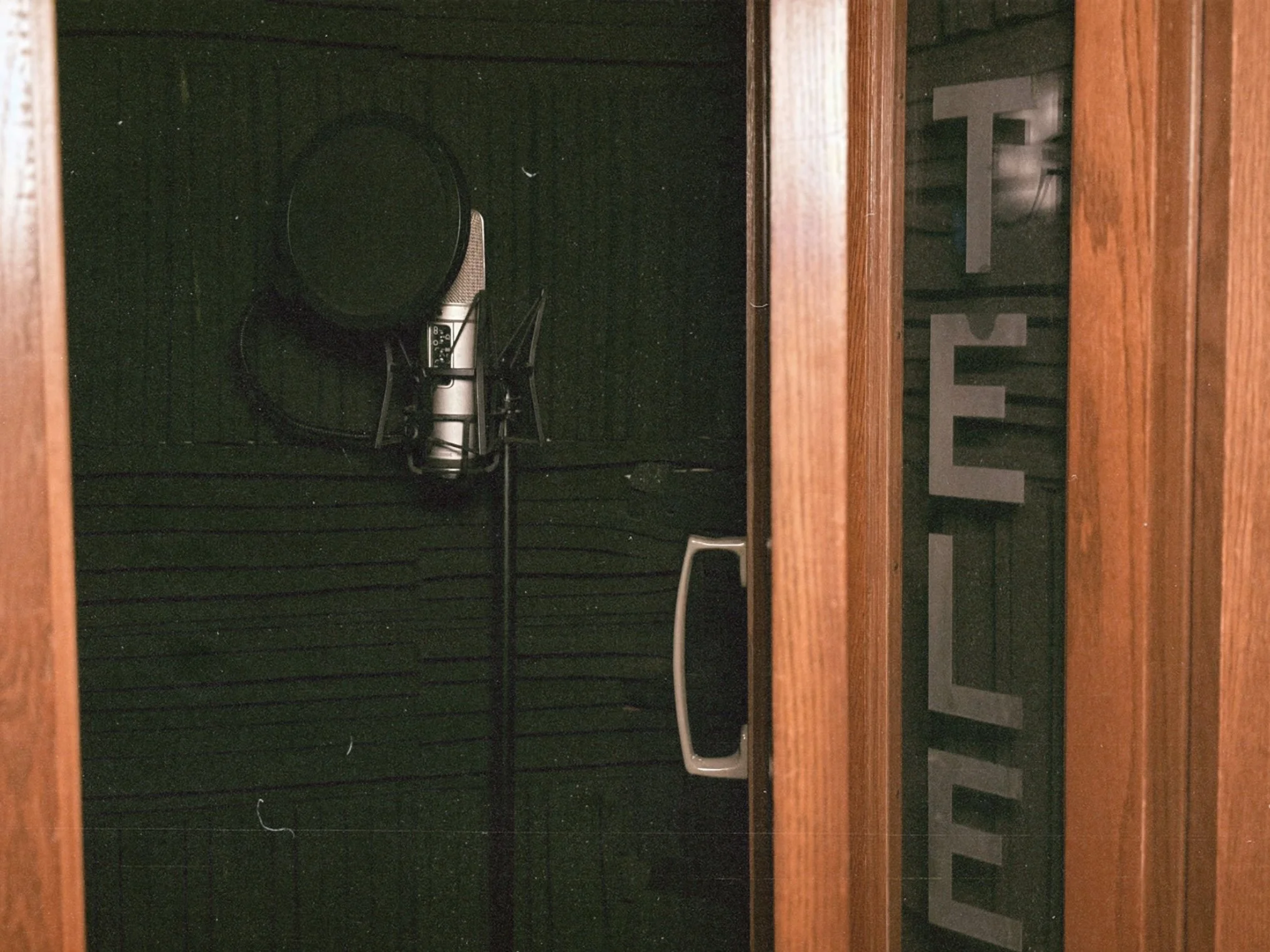 Interior view of a recording booth with black acoustic panels, containing a microphone, a chair, and a stand, seen through a window with the word 'VOICE' written on it.