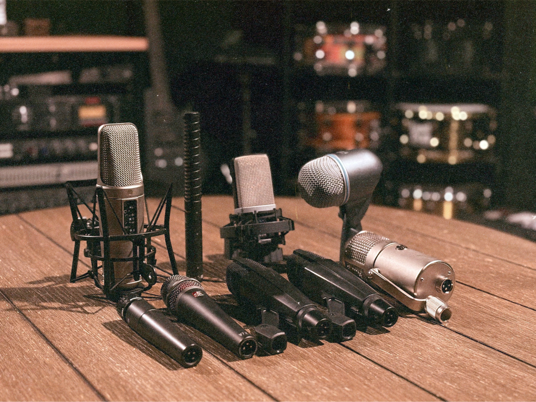 Collection of studio microphones on a wooden desk, with equipment racks and blurred background of audio studio.