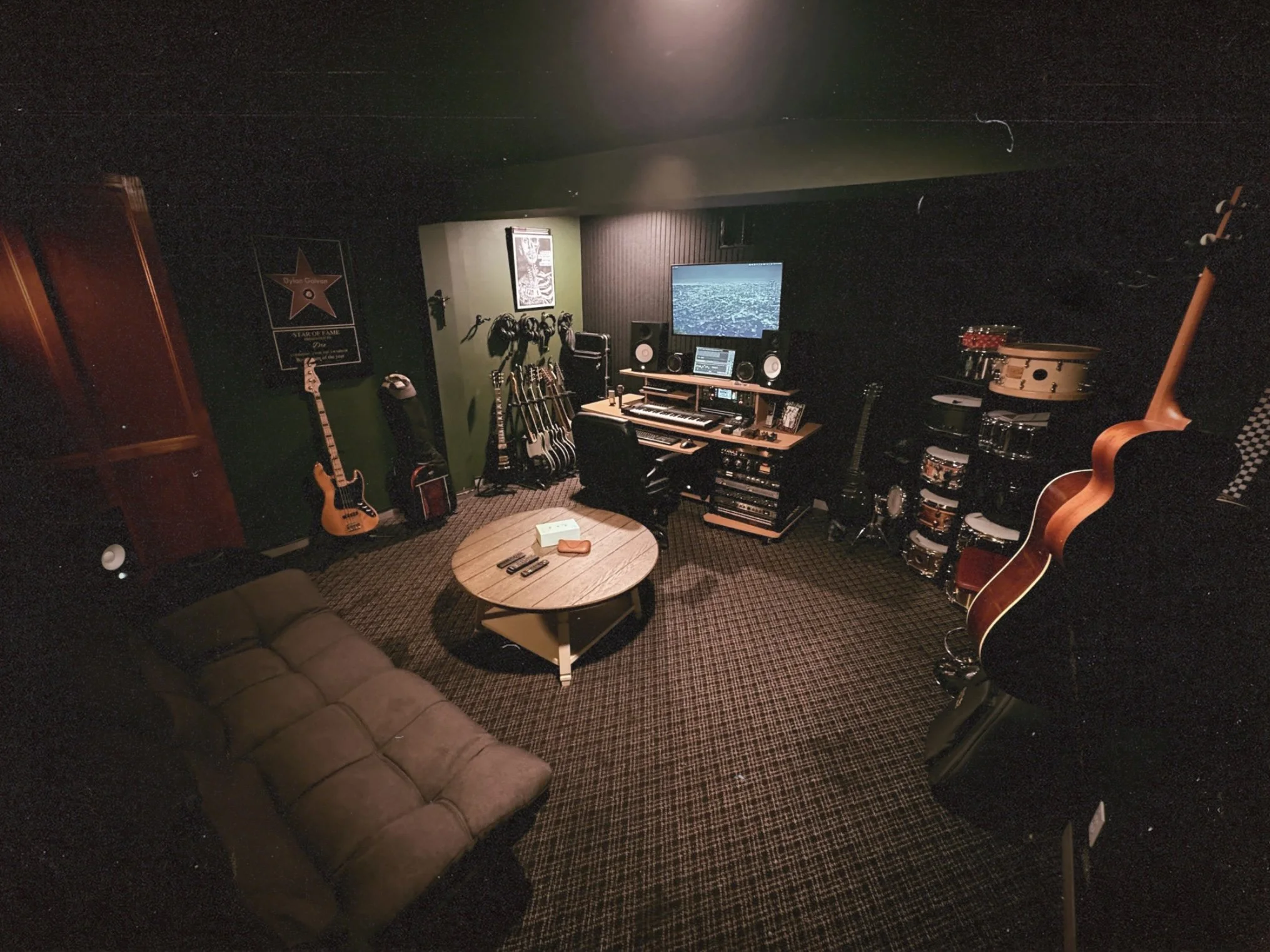 A music rehearsal room with guitars, drum set, and recording equipment, including a large screen monitor, and some musical accessories on a wooden coffee table.