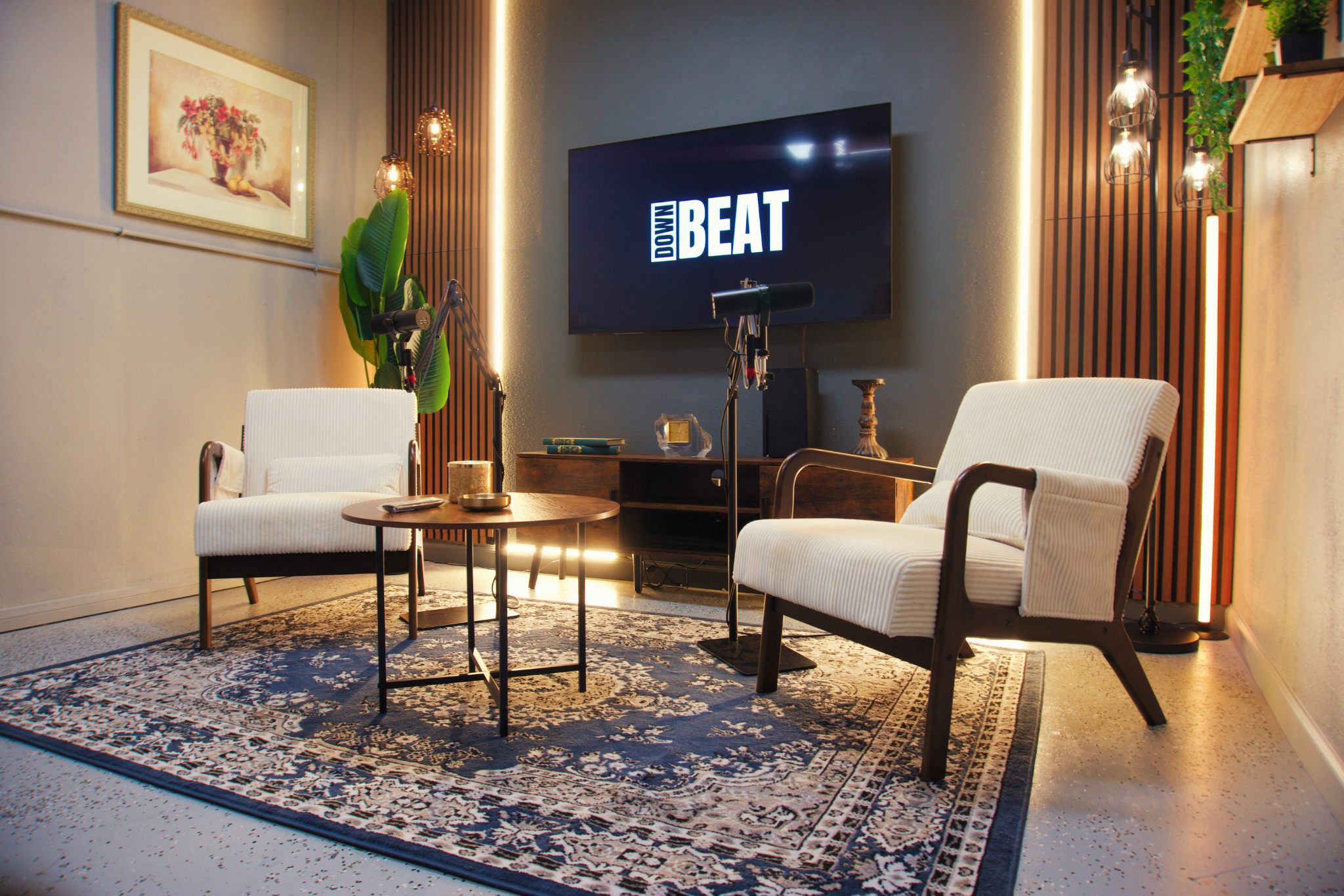 A cozy conference or meeting room with two white armchairs, a small round table, and a large wall-mounted screen displaying the words "DOWN BEAT." The room features a large decorative rug, wall art of a vase with flowers, vertical wooden slats, and modern hanging light fixtures with a warm glow.
