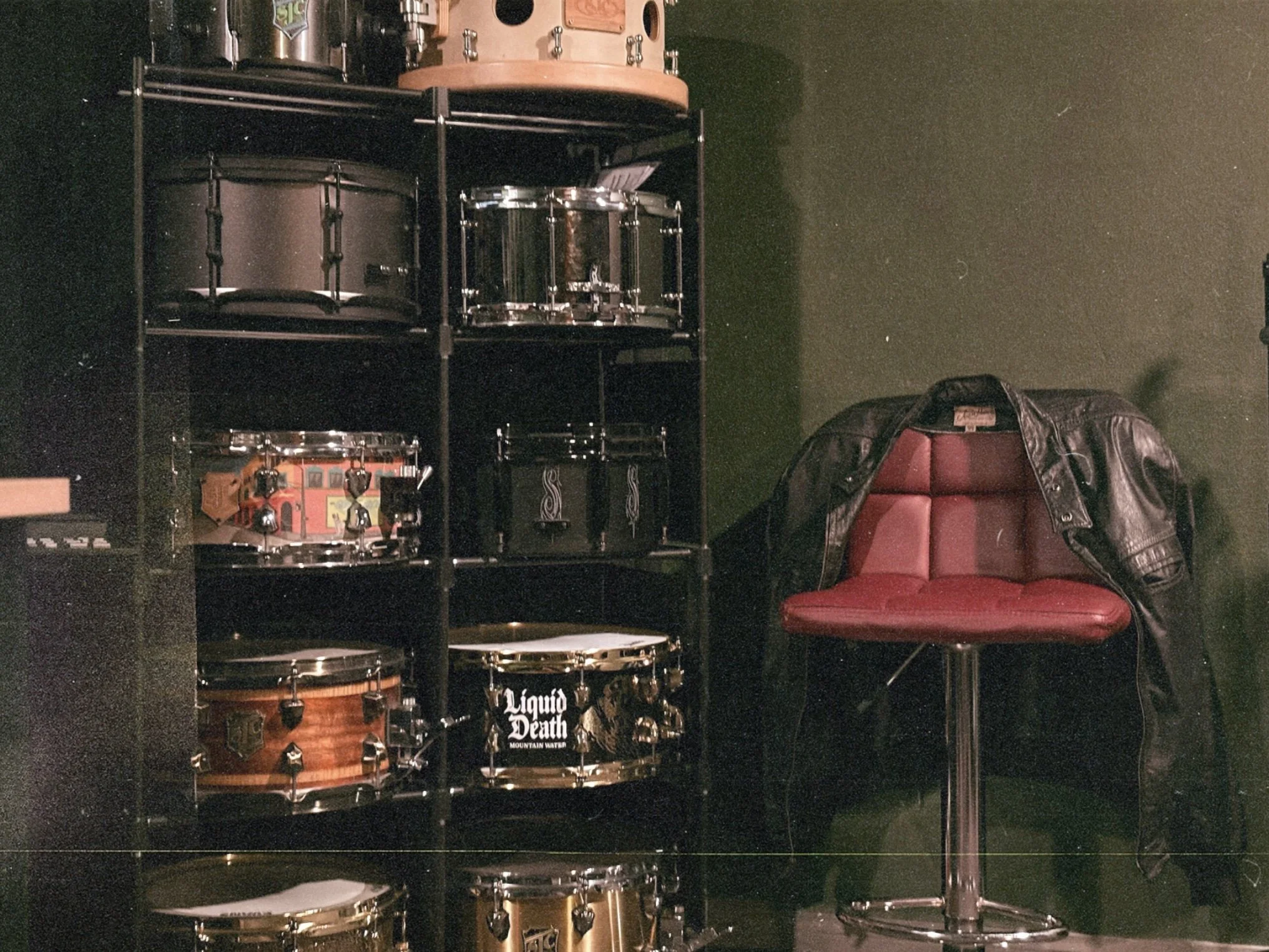 A black shelving unit with drums, pedals, and music equipment, next to a red and black leather chair and a black metal pole, in a dimly lit room with dark walls.